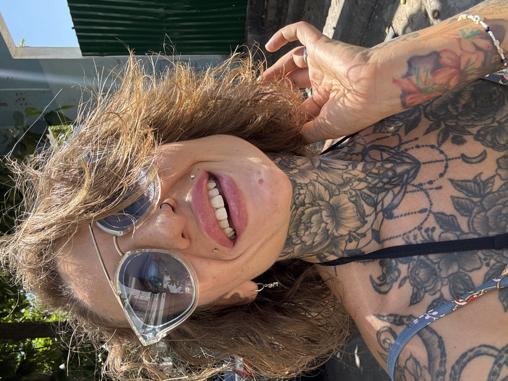 A woman is pictured outdoors, with her upper body and face taking up most of the frame. She has medium-length, wavy brown hair that appears windswept and is wearing large, clear-rimmed sunglasses. She has several piercings: one on her nose, one on her lip, and one on her cheek. Her mouth is open in a smile, revealing her teeth. The woman has extensive black ink tattoos covering her neck, chest, and extending onto her arm, featuring floral and other designs. She is wearing a dark-colored strap from her clothing, possibly a bikini top or dress. Her right hand is raised, touching her hair, and she is wearing a ring on her right index finger. On her wrist, there is a colorful tattoo of flowers and a beaded bracelet with skull charms. The background shows foliage from trees, a light-colored building with a window, and a dark green corrugated metal fence. The lighting suggests it is a sunny day. The provided location is Bac Nha Trang Ward, Vietnam.