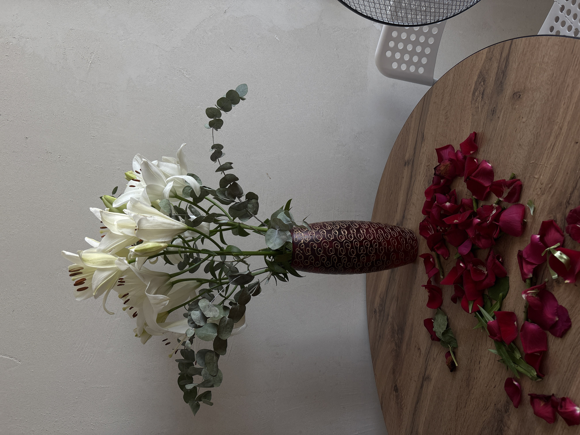 A still life arrangement is depicted on a circular wooden table, with a plain, textured light grey wall as the background. The table is positioned indoors, near a white plastic chair and part of a black fan.
In the center of the table sits a tall, slender, dark red vase adorned with a gold swirling pattern. It contains a bouquet of white lilies and eucalyptus leaves. The lilies are in full bloom, with their petals unfurled, revealing prominent dark stamen. The eucalyptus leaves are a muted green, providing a soft contrast to the white flowers.
Scattered across the surface of the table, in front of the vase, are numerous dark red rose petals and a few green stems. These petals appear to have fallen from a separate bouquet or have been intentionally strewn.
The lighting suggests an indoor environment, possibly with natural light coming from the side, casting soft shadows. The overall scene conveys a sense of aesthetic arrangement, with the fallen rose petals adding a touch of romantic or melancholic ambiance. The location of Rostov-on-Don, Russia is not visually indicated within the image.