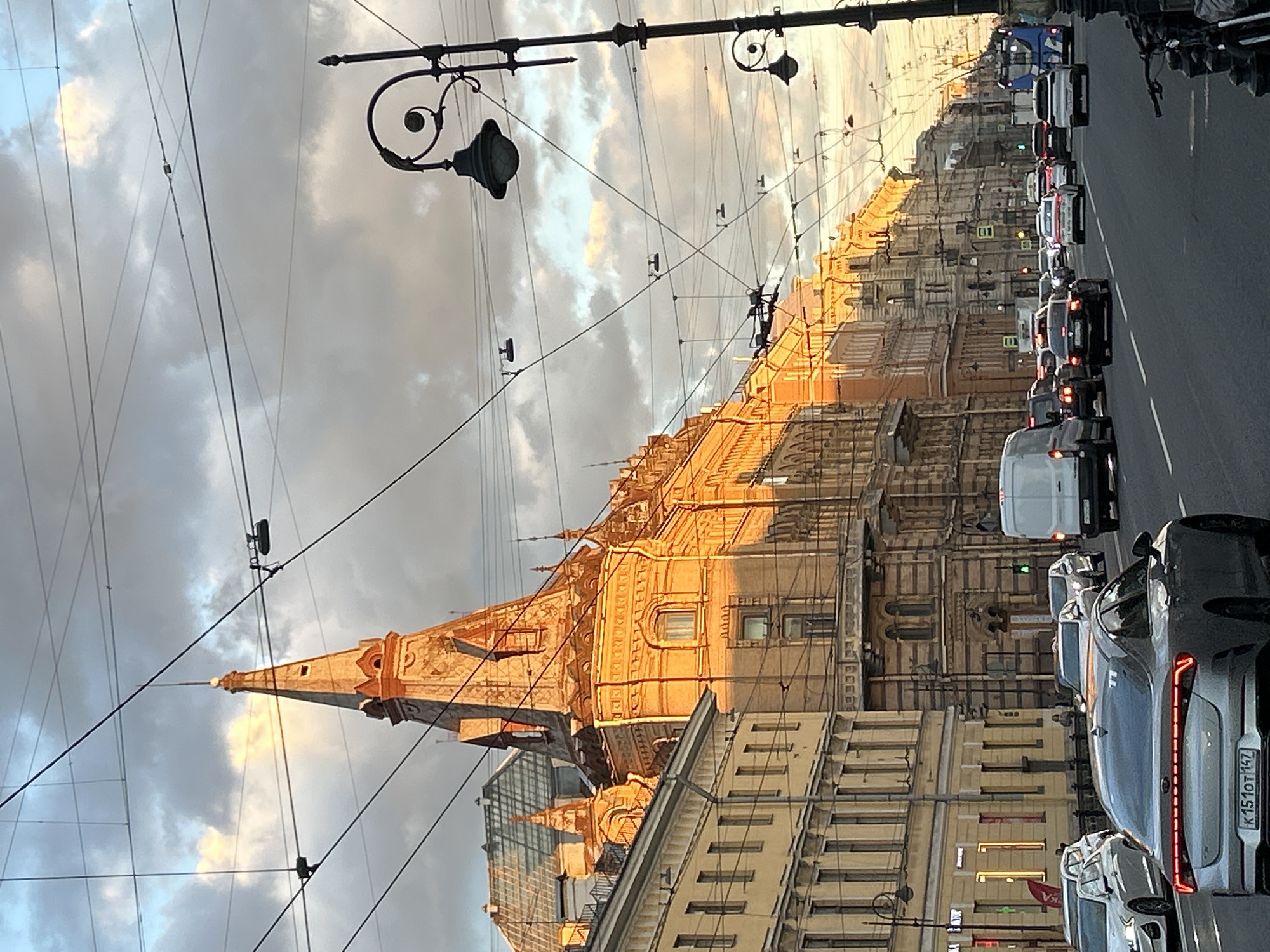 An eye-level shot captures a wide city street with several lanes of traffic during the day. The sun, low in the sky, casts a warm, golden light on the ornate, multi-story buildings lining the left side of the street, highlighting their architectural details and the prominent spire of one building. The street is filled with cars and a white van, all moving in the same direction. Overhead, a complex network of power lines and trolley wires crisscrosses the sky, obscuring parts of the sky and buildings. A decorative lamppost with a scrolled arm stands on the right side of the image. The sky is filled with dramatic, grey and white clouds, with patches of blue peeking through. The license plate on the silver car in the foreground reads "K151OT 147".