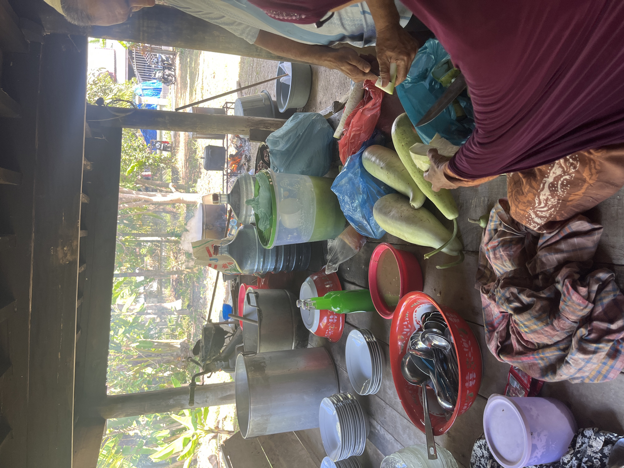 In Peurupok, Indonesia, a group of people are gathered around a rustic wooden table under a covered outdoor area, likely preparing a meal. The scene is dominated by cooking and food preparation items. Large metal pots, plastic water jugs, and stacks of plates are visible. Several people are actively involved in the preparation, with one person seen cutting vegetables and another seemingly preparing something in a red bowl. The environment appears to be outdoors, with lush greenery and trees visible in the background, suggesting a rural or semi-rural setting. The overall impression is one of community gathering and shared food preparation.