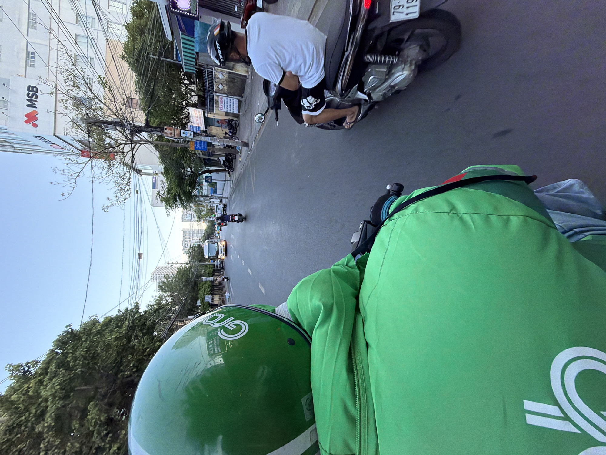 The image shows a first-person perspective of someone riding a motorcycle in Nha Trang, Vietnam. The rider is wearing a green helmet with a "Grab" logo and a green jacket, also with a logo. To the right, another person is riding a motorcycle, wearing a white t-shirt and black shorts. In the background, there are buildings, trees, power lines, and a bus. The road is paved and has lane markings. The scene appears to be during daylight, with a clear sky.