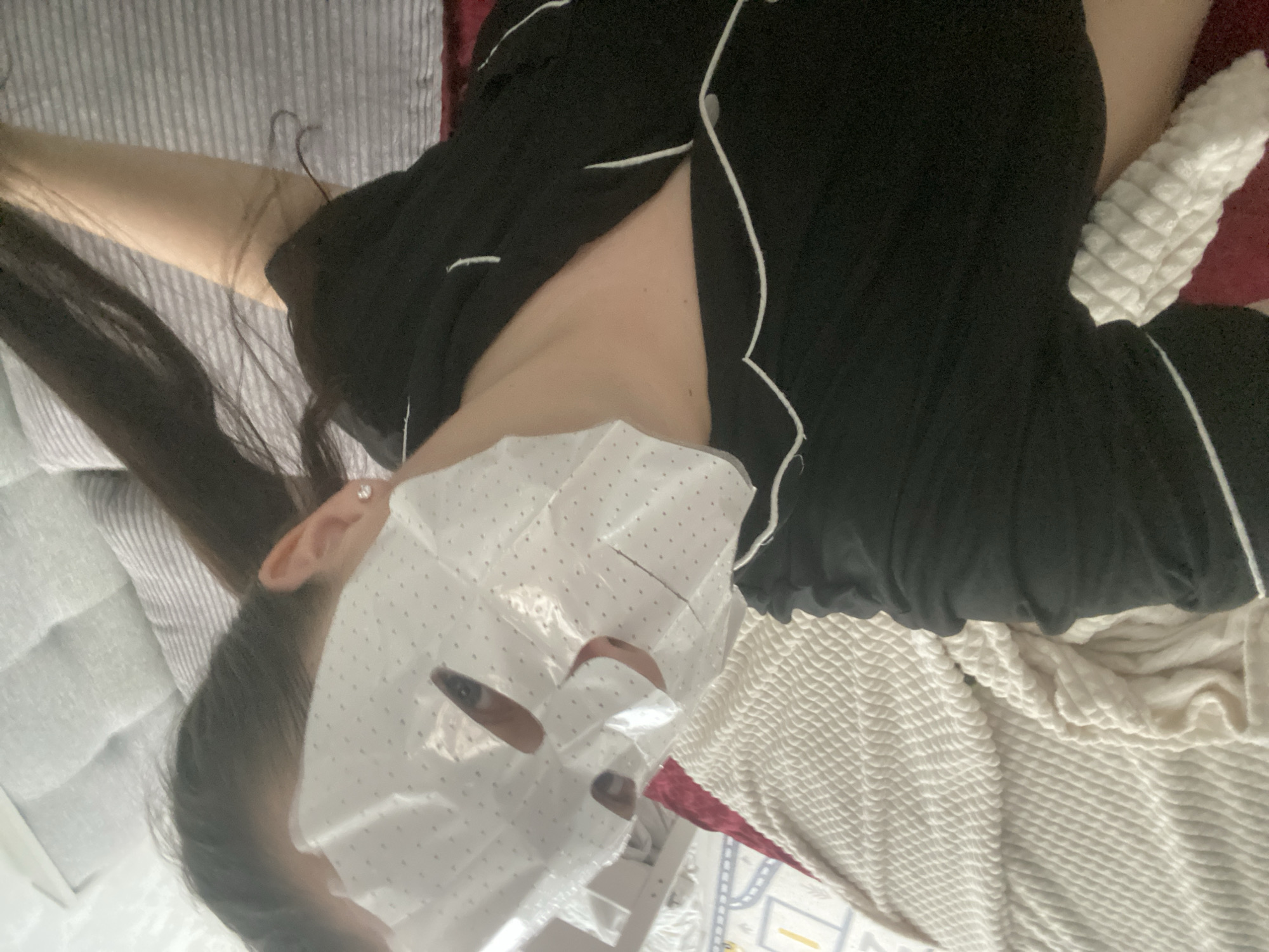 A person is lying on a bed with a facial sheet mask on. The person is wearing a black pajama set with white piping. The background includes a grey striped comforter, a white textured blanket, and a red pillow. There is a glimpse of a children's play area in the lower part of the image. The location is not discernible from the image.