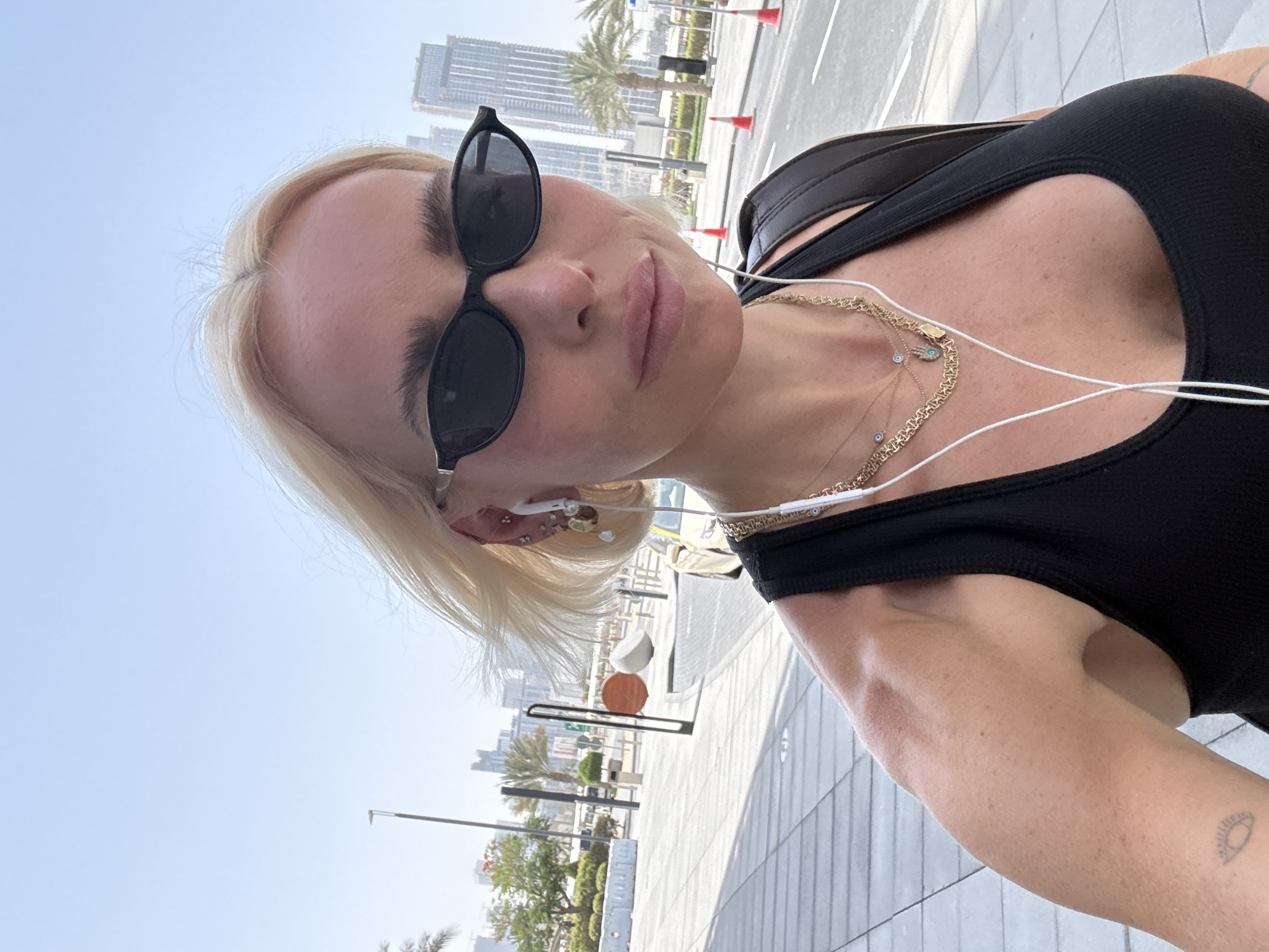 A medium-shot, selfie-style photograph shows a blonde woman wearing sunglasses and a black tank top outside in a city environment. She is facing the camera at a slightly downward angle. Her face is centered in the upper portion of the frame, and she appears to be wearing earphones. She has several necklaces around her neck. The woman's right arm is visible with a small tattoo on the inner part of her forearm.
In the background, there are tall buildings, palm trees, and traffic cones along a paved road and sidewalk, which indicates a city setting. Given the distinctive architecture and overall appearance, the location is identified as Dubai, United Arab Emirates. The sky is clear and bright, suggesting it is daytime. The sidewalk has a light gray, grid-like pattern. A partially visible structure with orange and white spheres is on the left.