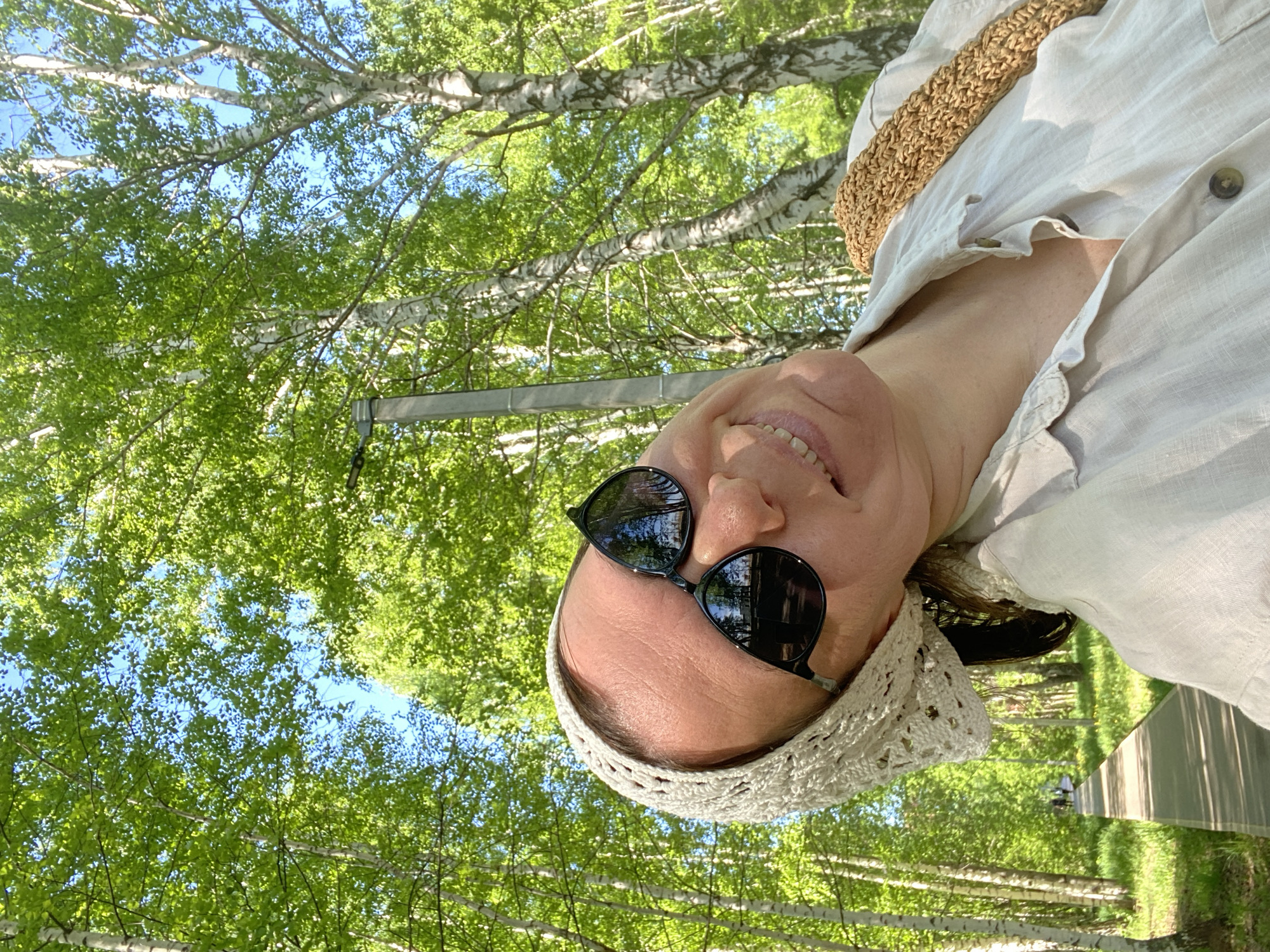 A selfie of a fair-skinned woman wearing sunglasses and a crocheted headband, angled upwards toward a canopy of green leaves and birch trees with white bark. The sunlight filters through the leaves. She is wearing a light-colored button-down shirt. The background includes a glimpse of a street or path. Given the specification of Chaikovsky, Russia, this image was presumably taken there. The woman appears to be smiling.