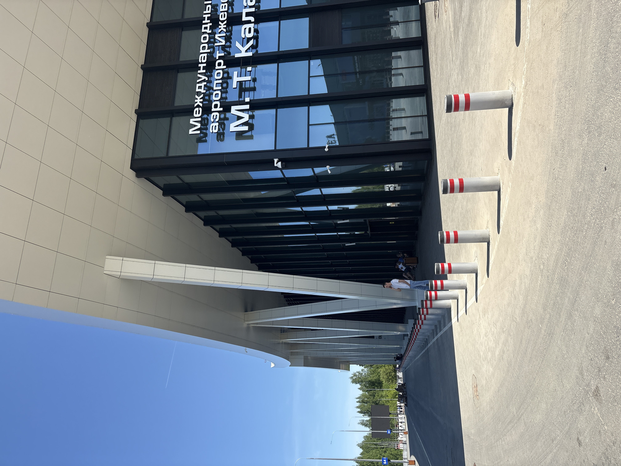 The image shows the exterior of the Izhevsk International Airport in Novoye Martyanovo, Russia. The building has a modern design with a glass facade and a large overhang supported by white beams. The name of the airport is displayed in Russian on the glass. A man in a white shirt and jeans is seen standing near the entrance. Several white and red cylindrical bollards line the sidewalk in front of the building. The sky is blue and the environment appears to be sunny.