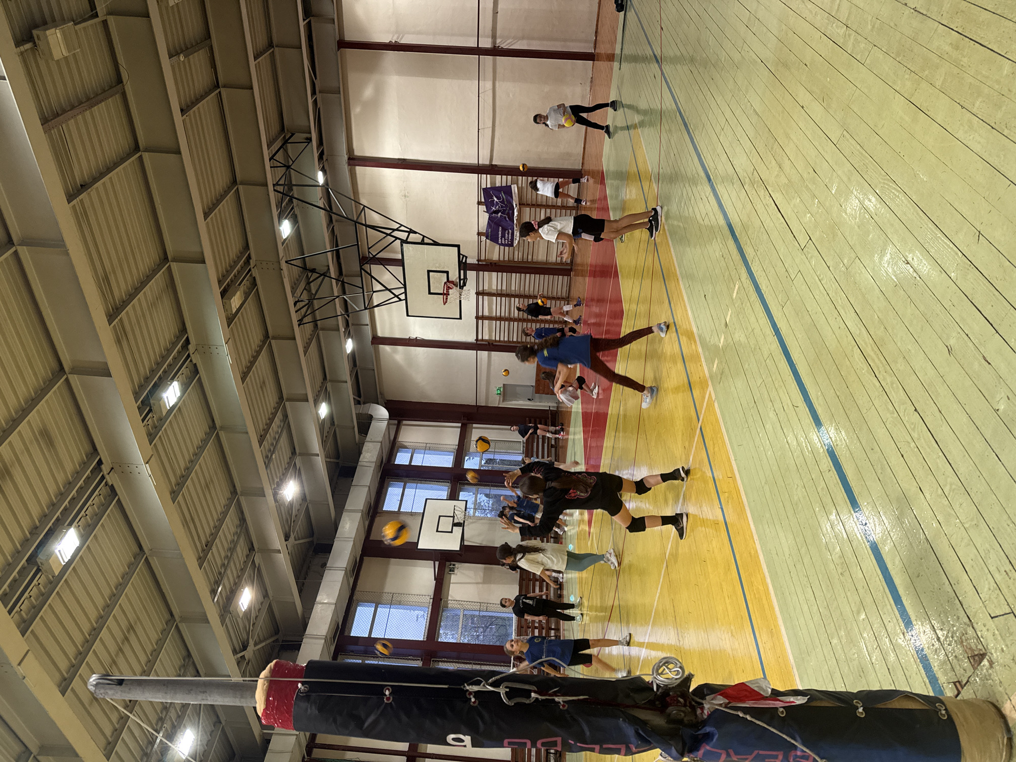 This media file captures an active indoor volleyball practice or training session in a gymnasium, likely located in Sofia, Bulgaria. The image is taken from a low perspective, looking across the court towards the far wall, with a volleyball net pole prominently positioned in the immediate foreground on the left.
The setting is a large sports hall characterized by a high, industrial-style ceiling featuring exposed metal beams, trusses, and corrugated panels. Numerous rectangular artificial light fixtures are embedded in the ceiling structure, providing bright illumination. Windows line the upper sections of the walls, allowing some natural light to filter in, suggesting it might be late afternoon or evening. The court floor is made of polished wood, segmented into distinct light yellow and a paler greenish-yellow areas, separated by thin blue lines, typical for multi-sport gymnasiums. Basketball hoops with backboards are also visible along the walls, indicating the facility's versatility.
Multiple young individuals, appearing to be teenagers, are scattered across the court, actively engaged in the session. They are dressed in athletic attire including t-shirts, shorts, leggings, and sneakers. Several yellow and blue volleyballs are present; some are in the air, some are being held by players, and others are on the court floor, suggesting various drills or a game in progress.
Notable details include a banner displayed on the far wall with a purple background, featuring stylized figures that resemble gymnasts or dancers. Text on the banner is partially visible in Bulgarian, including "Гимнастически спортове" (Gymnastic sports) and likely "Федерация" (Federation) below it. In the foreground, the dark blue padded cover of the volleyball net pole has partially visible white text that appears to read "BEACH". The overall atmosphere is one of focused activity and athletic training.