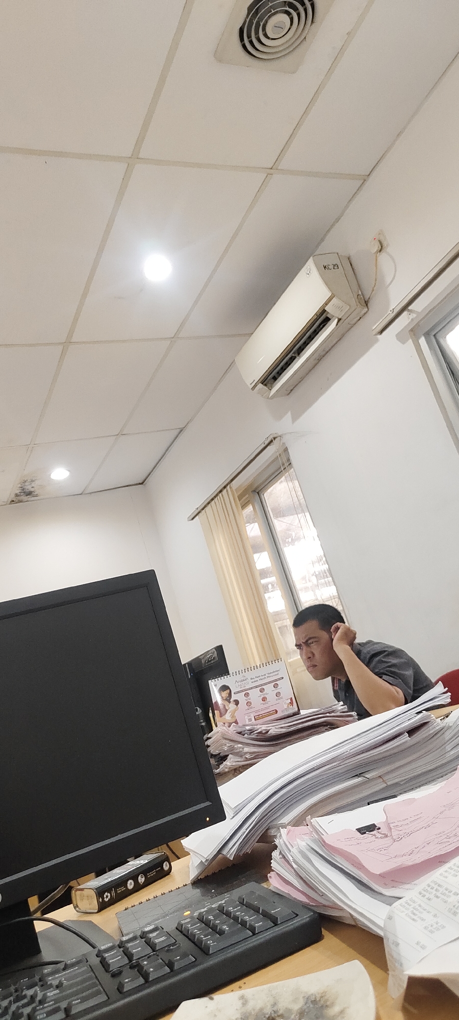 This image depicts an indoor office environment, possibly a small business or personal workspace, in the city of Nusa Indah, Indonesia. The scene is dominated by a desk cluttered with stacks of papers and documents. In the foreground, a black computer monitor is visible, with a keyboard positioned in front of it. A cylindrical object with various symbols is placed on the desk behind the keyboard.
To the right of the desk, a man is seated, looking intently at the computer screen. He has dark hair and is wearing a dark t-shirt. His facial expression suggests concentration or perhaps frustration, as he is seen resting his head on his hand. Behind the man, a window with beige curtains allows natural light to enter the room.
The office ceiling is made of white tiled panels, with a circular air vent and a light fixture visible. An air conditioning unit is mounted high on the white wall. The overall impression is one of a busy, somewhat disorganized workspace.