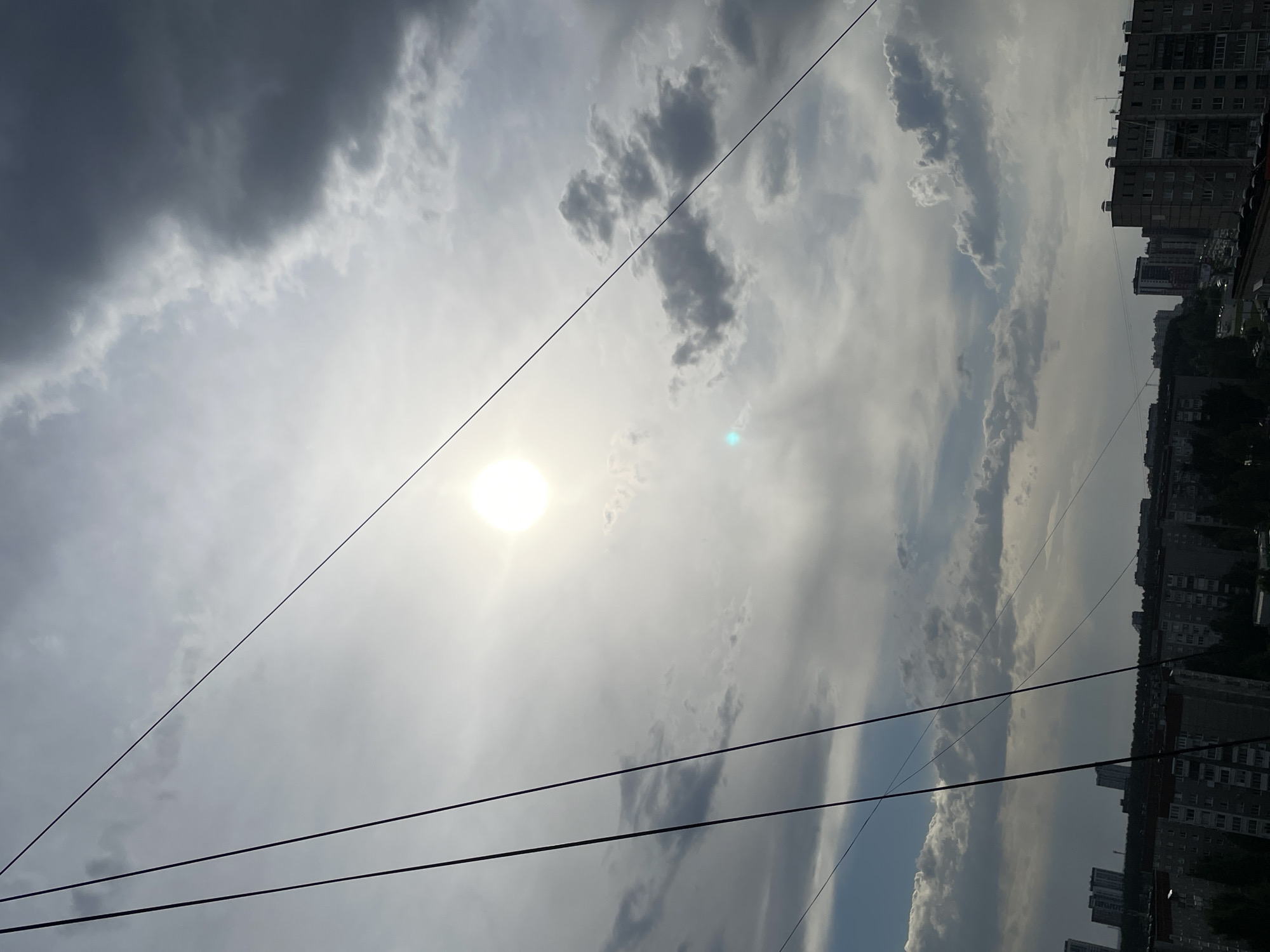 The image depicts a daytime sky with scattered clouds and the sun partially obscured. Power lines are visible in the foreground, and a section of a city with numerous buildings is on the right side of the frame. The location is Izhevsk, Russia.