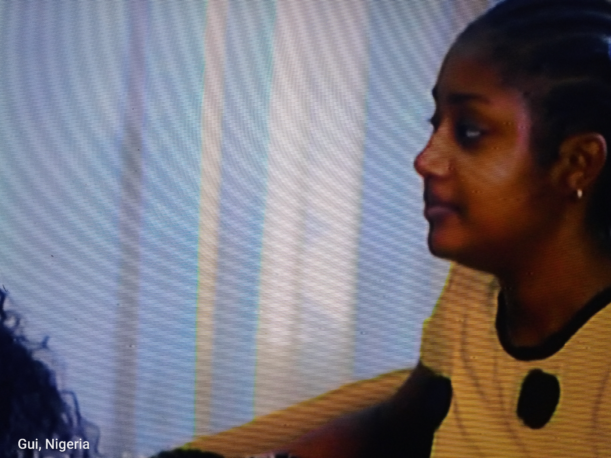 The image shows a side profile of a Black woman with braided hair and a small hoop earring. She is wearing a yellow top with a black circular design. The background is out of focus and appears to be a wall with vertical stripes in shades of blue and white. Text at the bottom of the image reads "Gui, Nigeria."