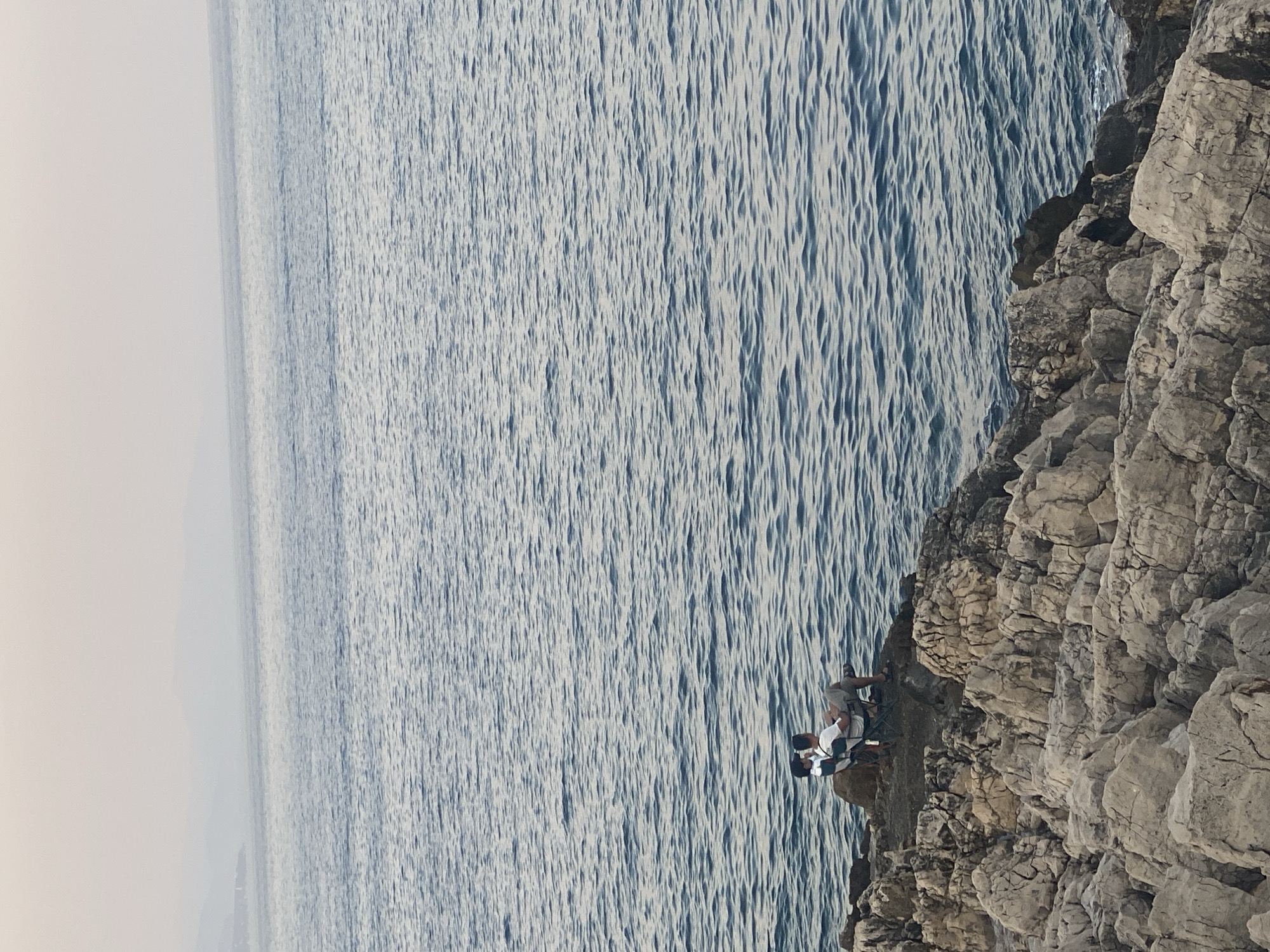 Two individuals are seated on folding chairs on a rocky outcrop overlooking the sea. The person on the left, further from the camera, is wearing a dark shirt and appears to be a child. The person on the right, closer to the camera, is an adult male wearing a white t-shirt and shorts. Both individuals are facing the sea. The sea itself is a deep blue with visible ripples. The rocky outcrop is composed of light-colored, textured rocks. In the far distance, hazy landmasses and a pale sky are visible. This scene is located in Antibes, France.