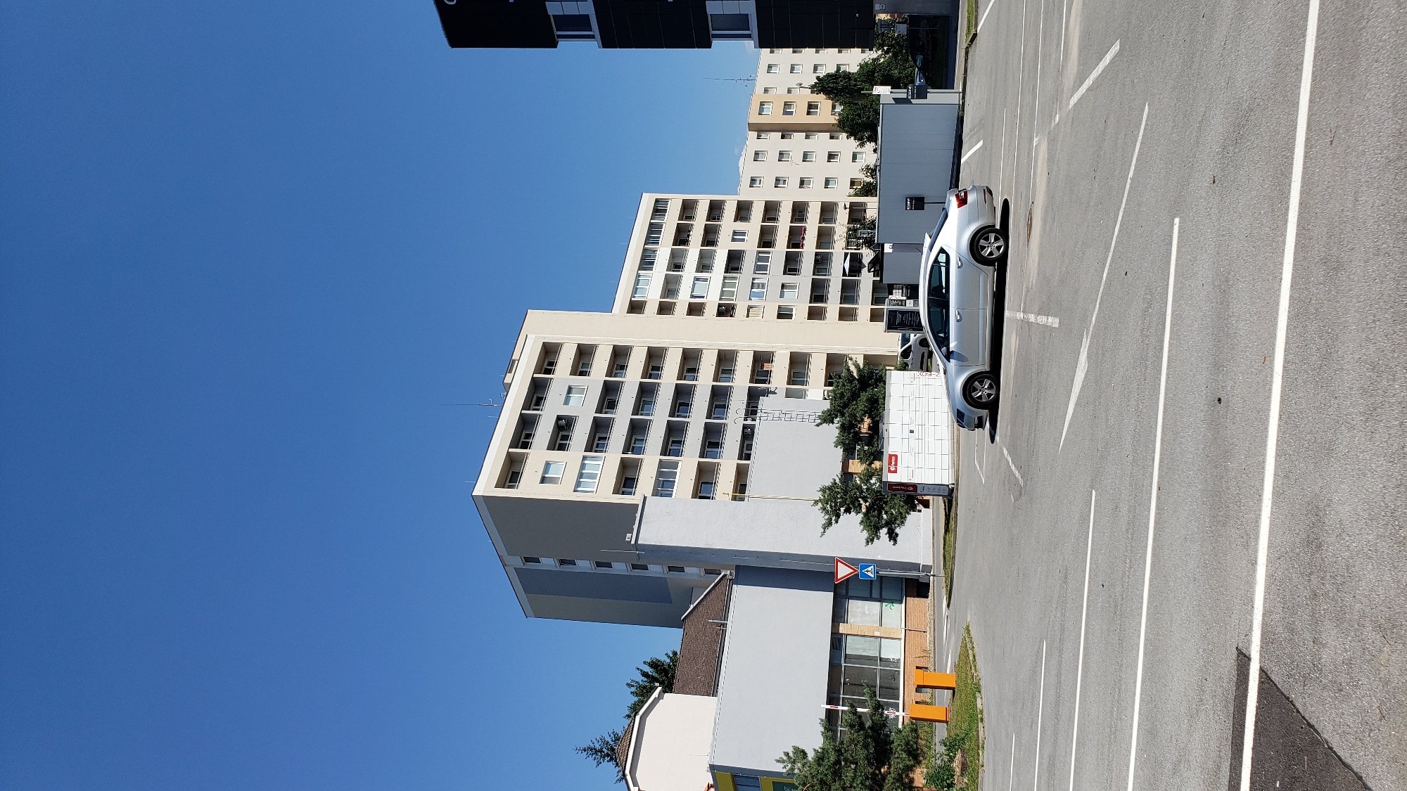 This is a photograph of a residential area on a sunny day, likely in Prešov, Slovakia. The scene is dominated by several multi-story apartment buildings, one of which is a prominent, light-beige building with numerous balconies. Another building in the foreground is gray with a light beige upper section. There are also glimpses of darker buildings in the background, suggesting a densely populated urban environment.
In the foreground, a silver car is parked in a marked parking lot. The parking lot is paved with asphalt and has white painted lines indicating parking spaces, as well as directional arrows. Next to the parking lot and buildings, there are some trees and greenery.
On one of the gray buildings, there is a white structure that appears to be a parcel locker system, with several compartments. There is also a red sign with white lettering on this structure. A blue and white triangular traffic sign with a red border is visible on a pole near the buildings, indicating a warning.
The sky is a clear, bright blue, indicating a sunny day with good weather. The lighting suggests it is daytime, likely late morning or early afternoon given the shadows. The overall impression is one of a typical urban residential setting. There is no visible text that can be clearly deciphered.