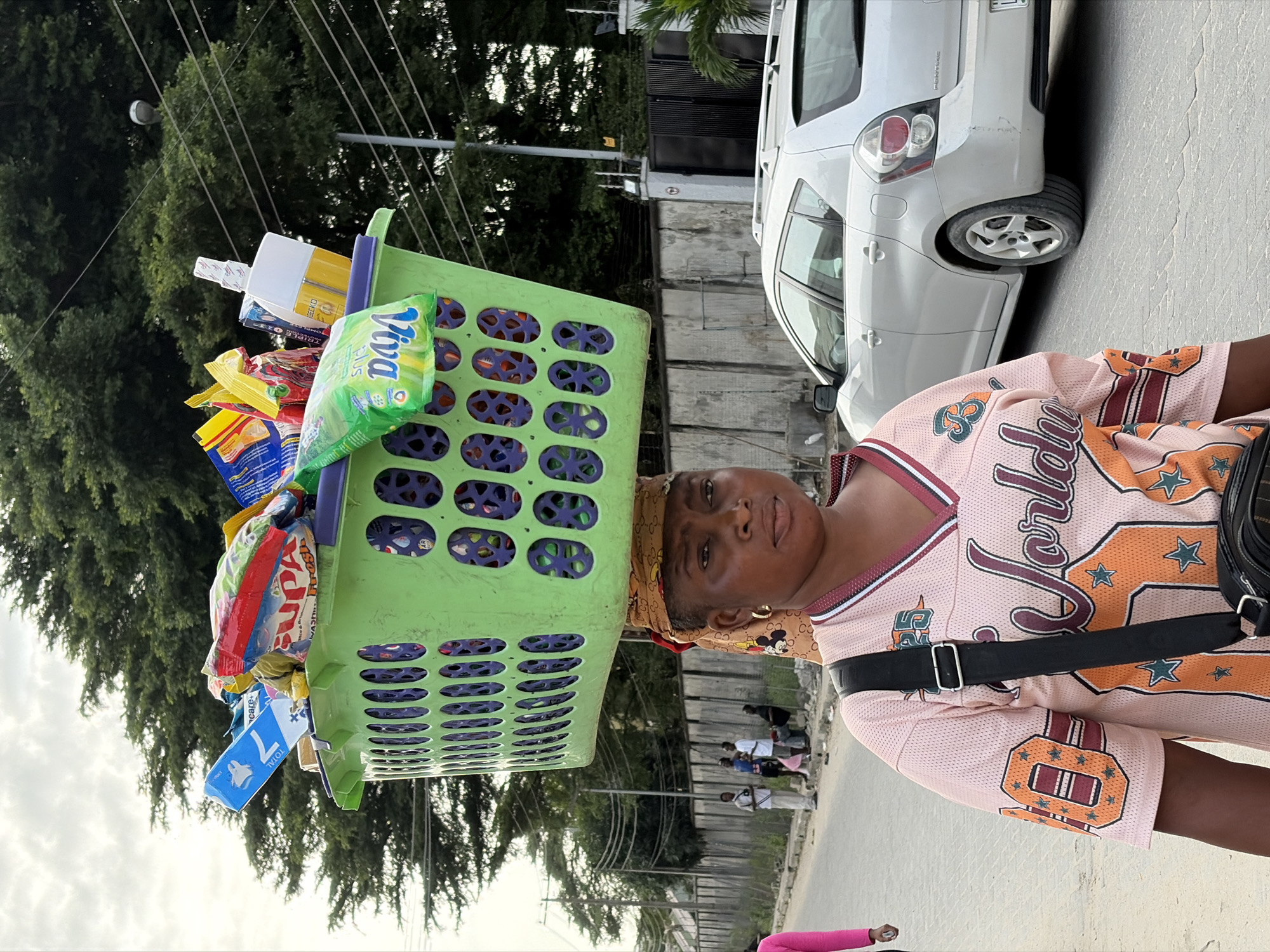 In Igboefon, Nigeria, a woman carries a large green plastic basket filled with various packaged goods on her head. She wears a pink jersey-style shirt with orange and white lettering and designs, and a patterned headwrap. A black strap from a bag is visible across her chest. Behind her, a light-colored car is parked on a paved road. In the background, several other individuals are standing near a fence, with lush green trees and power lines overhead. The sky is overcast.