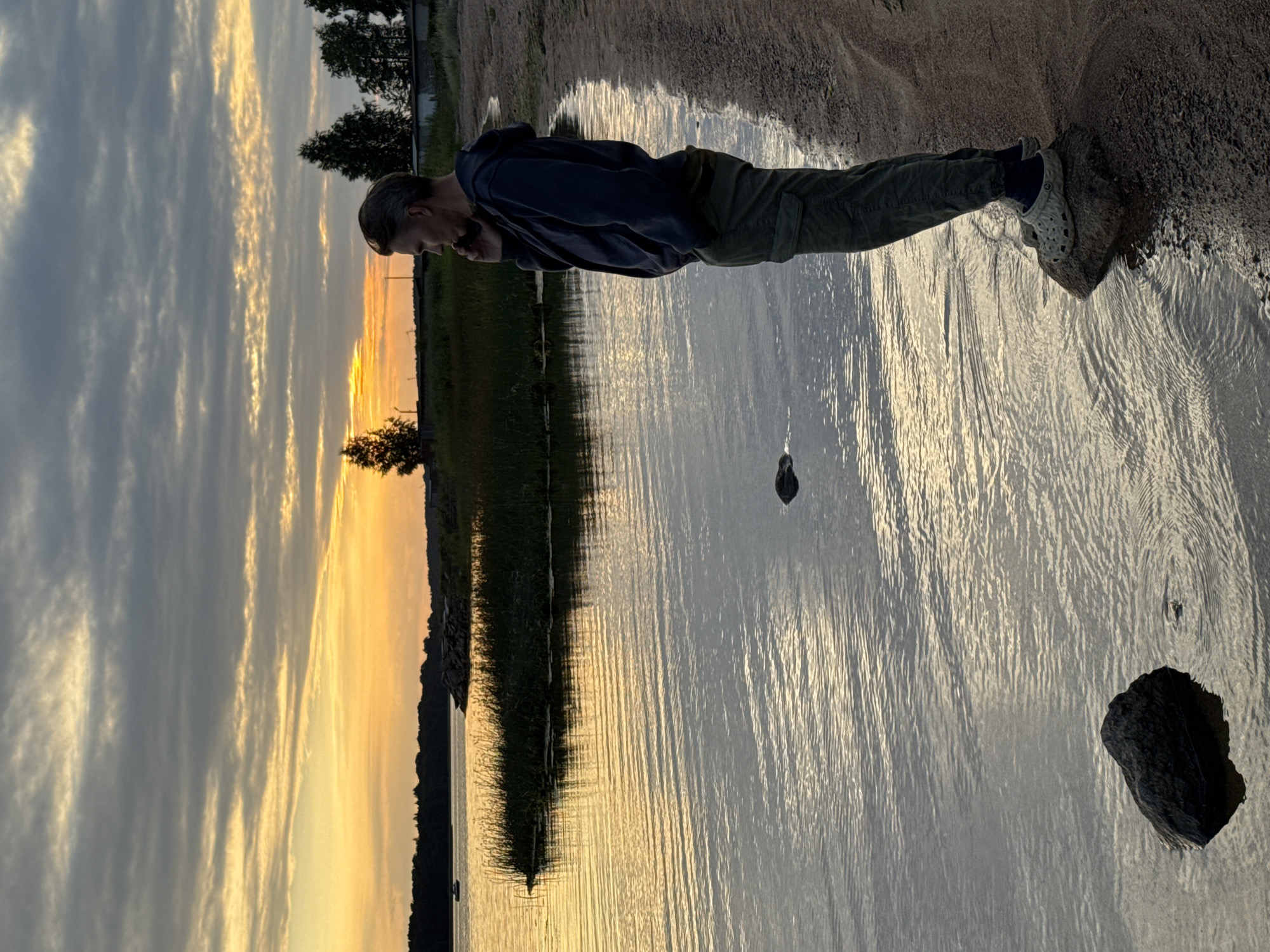 The image depicts a man standing on a rock at the edge of a body of water, possibly a bay or lake, during sunset. The man is wearing a dark hoodie and green cargo pants, with his right hand near his face, suggesting he might be on a phone call. He is facing away from the camera, looking towards the horizon. The sky is filled with dramatic clouds, with streaks of orange and yellow from the setting sun illuminating the scene. The water is calm with visible ripples, reflecting the colors of the sky. A patch of tall grass and reeds is visible along the shoreline in the middle ground, and further in the distance, trees and buildings can be seen. The shoreline is sandy and appears to be a natural setting. The overall impression is one of quiet contemplation or a moment of connection amidst a natural landscape at dusk. While the prompt mentions the city of Glebychevo, Russia, this location is not explicitly discernible from visual cues within the image itself.