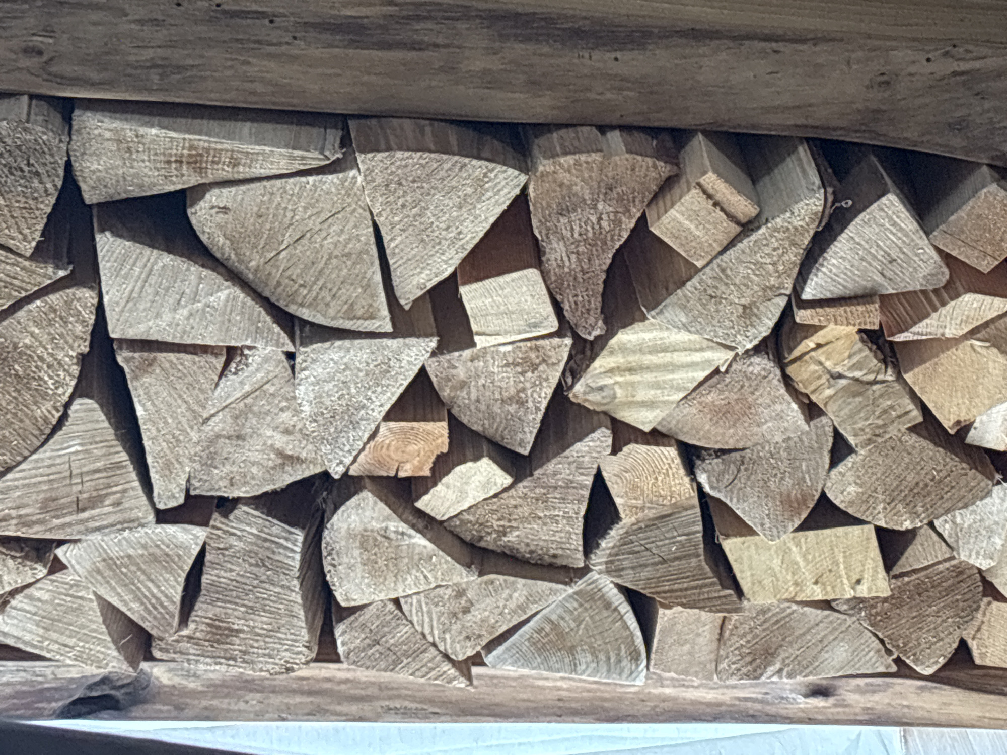 The image displays a close-up, eye-level view of a stack of firewood. The firewood is composed of many small, irregularly shaped pieces of wood, predominantly light brown and tan in color, with visible wood grain and some showing cut ends. The pieces are tightly packed together, creating a textured surface. To the left of the firewood, a light blue fabric or tarp is visible, partially obscuring the edge of the stack. To the right, a vertical wooden beam or post is present, its surface weathered and showing a darker wood tone. The lighting suggests an indoor or shaded outdoor environment, with shadows cast across the wood pieces. There are no people or animals present. This scene is not identifiable as being in Zvenigorod, Russia, or any specific city.