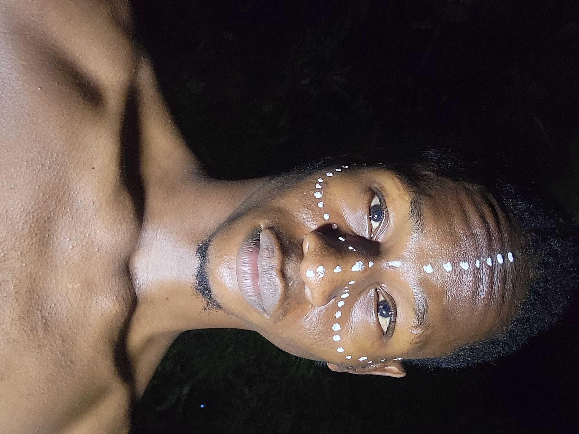 A close-up, dimly lit image depicts a shirtless Black man with white dot-patterned markings on his face, specifically on his forehead, nose, and cheeks. He has short, dark hair and a light beard. The background is a dark, indistinct mix of green foliage and black. The angle of the shot appears to be low, tilting upwards, which gives a sense of the man looking down on the viewer. The location is specified as Nkpor, Nigeria.