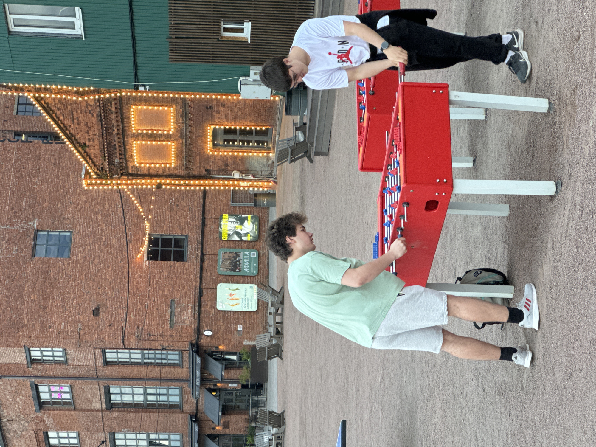 Two young males are playing foosball in an outdoor public area. The male on the left is wearing a light green t-shirt, light grey shorts, and white sneakers with red accents. The male on the right is wearing a white t-shirt with a red Jumpman logo, black pants, black sneakers, and glasses. Both are actively engaged in playing the game. The foosball table is bright red with white legs. In the background, there are brick buildings with large windows and some are decorated with string lights. There are also outdoor seating areas with wooden chairs and tables, and some posters are displayed on the brick walls. The ground is covered in gravel. The overall environment suggests a recreational area or a public space within a city. The posters and the architectural style of the buildings indicate the location is likely within Russia, potentially Saint Petersburg, given the style of the brickwork and the presence of Cyrillic text on some of the posters in the background.