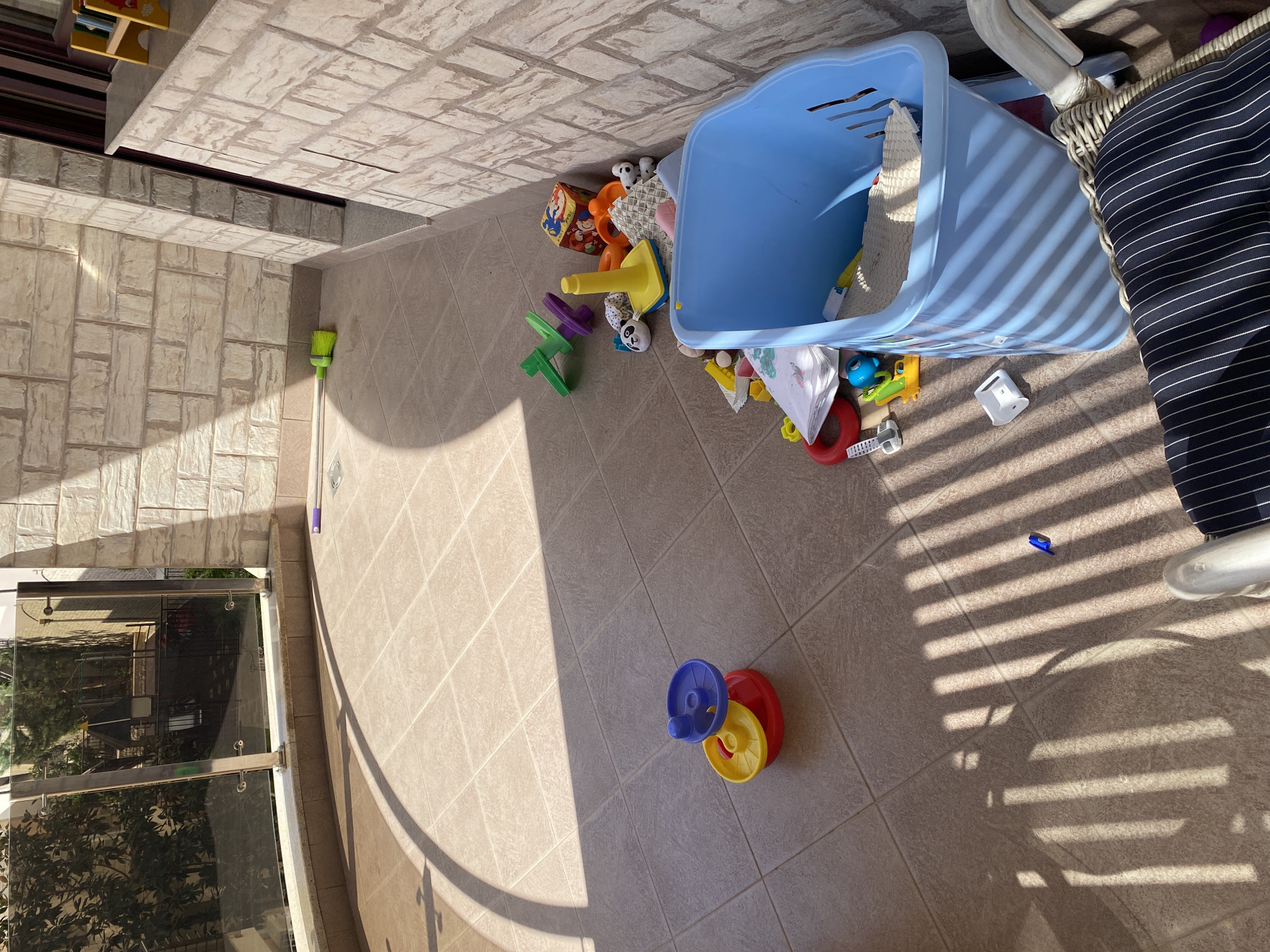 This image, taken in Bečići, Montenegro, captures a sunlit outdoor balcony or patio area with children's toys scattered across the tiled floor. No people are visible in the scene.
The primary subjects are a collection of colorful plastic toys and a large blue plastic bin, which appears to have been overturned or emptied, spilling its contents. Among the toys are various stacking blocks, a yellow plastic cone, a green and purple spinning toy, a red ring, a blue toy vehicle, and small white plastic pieces. Also visible are a few soft toys, some with panda-like patterns. Further away from the main cluster of toys, a set of red, yellow, and blue stacking or spinning wheels sits on the floor. A bright green broom with a long handle leans against the wall in the upper left section of the image.
The setting is characterized by light-colored, square floor tiles and a wall constructed from larger, textured rectangular stones. To the far left, a glass balcony railing offers a reflection of green foliage and distant structures, indicating an outdoor view. Part of a striped cushion or chair is visible on the right, next to the blue bin.
The time of day is evident from the strong, well-defined shadows, likely cast by blinds or a slatted overhead structure, which stretch diagonally across the floor and the blue bin. These long, distinct shadows suggest either mid-morning or late afternoon on a very sunny and clear day. There is no visible text in the image. The scene portrays a moment of disarray, typical after a period of children's play in a domestic outdoor space.