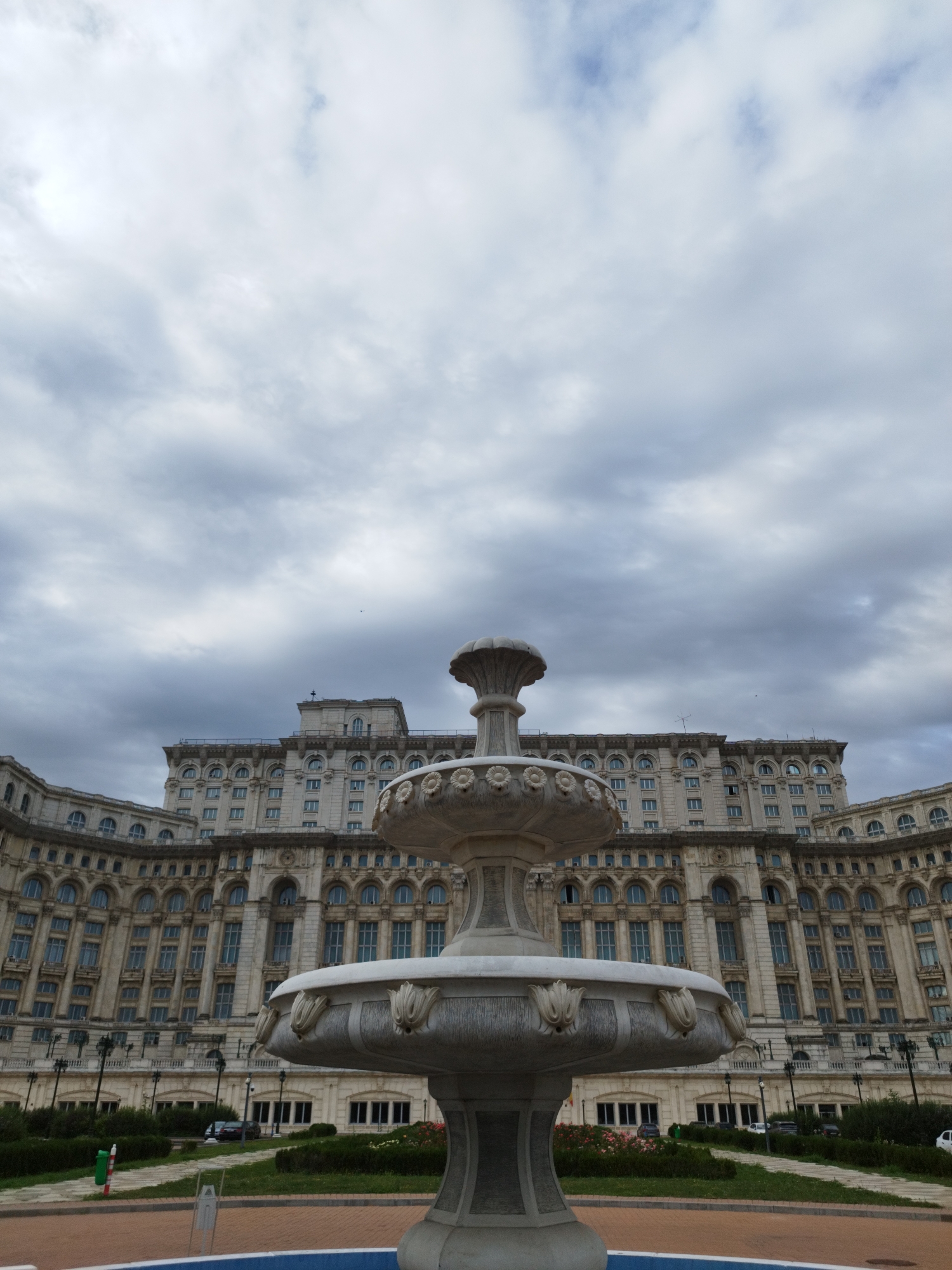 A low-angle, wide shot captures a monumental neoclassical building in Bucharest, Romania, under a cloudy sky. In the foreground, a large, ornate stone fountain dominates the frame, its tiers and decorative elements rendered in a light color. Beyond the fountain, a vast expanse of the building's facade stretches across the midground. The building features rows of arched windows, a pedimented central section, and symmetrical wings. The facade is detailed with columns, cornices, and decorative reliefs. A manicured lawn with hedges and lamp posts separates the fountain from the building. A few cars are visible parked in front of the building. The overall impression is one of grandeur and architectural scale, set against a somewhat overcast atmosphere.