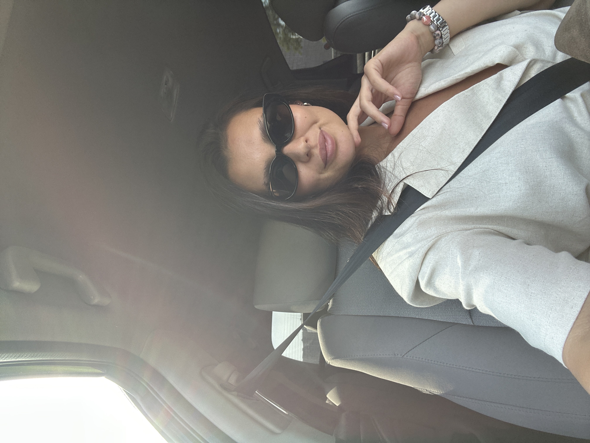 A woman with dark hair and sunglasses is in the passenger seat of a car. She is wearing a light-colored blazer and a seatbelt. The sun is shining through the window, creating a lens flare. The interior of the car is visible, including the seats and a handle on the ceiling. The image is taken from a selfie perspective. The location is not discernible from the image and there is no information to indicate that it is the city of Perm, Russia.