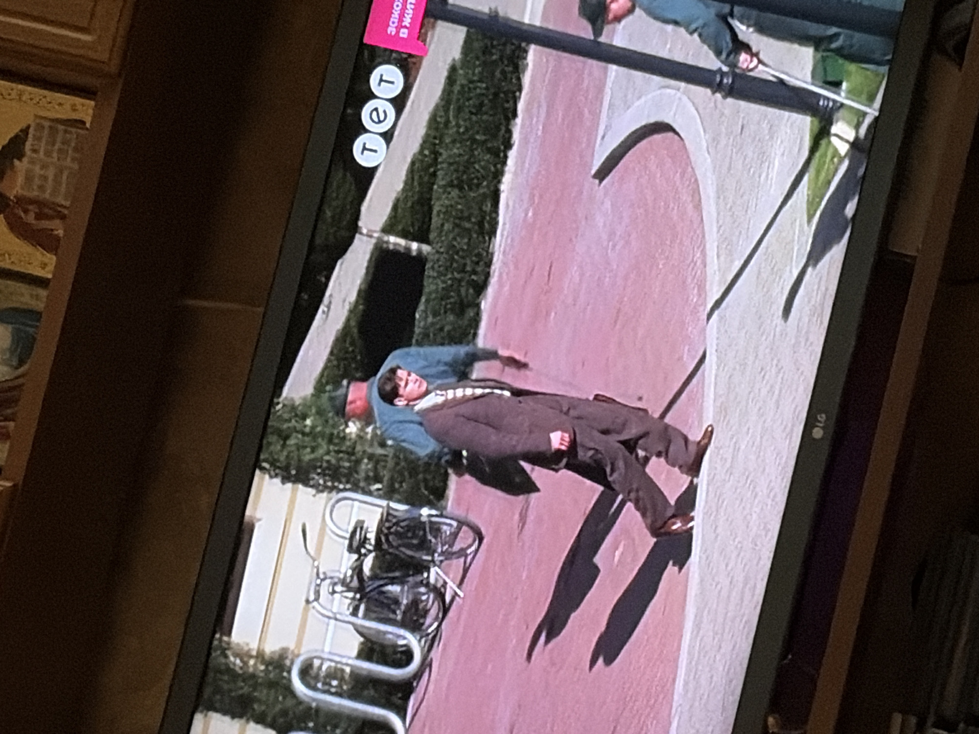 The image shows a screen displaying a scene with a man in a brown suit walking across a pink paved area. He is casting a long shadow. To his left, a bicycle is parked next to a silver rack. In the background, a person in a blue shirt and green hat is partially visible, and further to the right, another person wearing a green uniform and a cap is holding a white object. There are also decorative elements and logos visible on the screen, including "tet" and a pink sign with text in Cyrillic that appears to say "Закон жизни". The overall scene appears to be an outdoor setting, possibly in a park or public space. The location is identified as Osolynka, Ukraine.