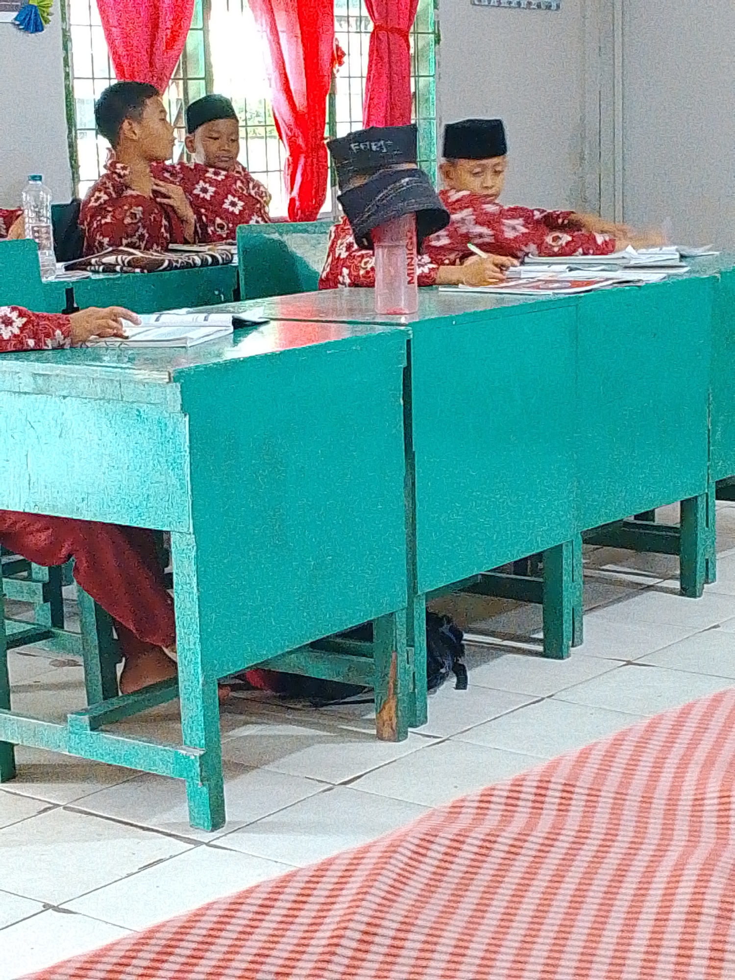 The image depicts an indoor scene, likely a classroom in Tembung, Indonesia, with several young boys wearing traditional red and white patterned shirts and black caps seated at green desks. In the foreground, a boy is seen from the waist down, his legs beneath the desk, wearing a red garment. To his left, another boy is partially visible, also in a red and white patterned shirt. Further back, two boys are seated at desks, engrossed in their studies or activities. One boy, wearing a black cap, is writing or drawing on papers, with a pink water bottle labeled "MINIGO" beside him. The room has red curtains and a window, suggesting it's daytime. The floor is tiled, and a red and white striped mat is visible in the lower right corner. The overall impression is of a quiet learning environment.