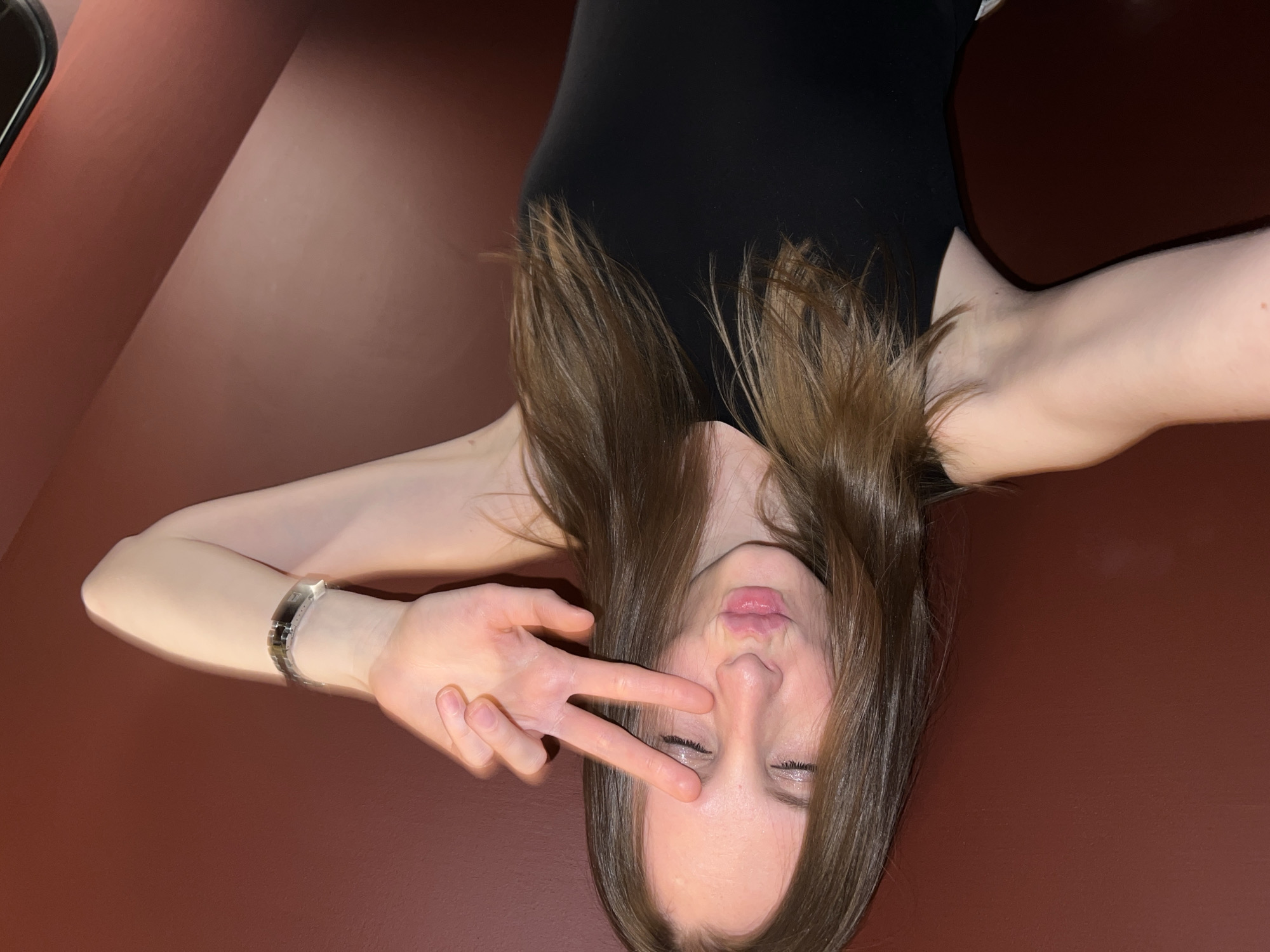A young woman with long, straight brown hair is depicted in a portrait, rotated 90 degrees clockwise from a typical upright orientation. She is wearing a simple black top or dress. Her right hand is raised to her face, forming a "peace" sign with her index and middle fingers. Her eyes are closed or squinted, and her lips are pursed in a playful expression. A metallic watch is visible on her left wrist, which is bent, with her left arm extending towards the top of the frame. The background is a plain, dark reddish-brown surface, likely an indoor wall, with subtle variations in lighting creating a diagonal tonal shift. The overall mood is casual and playful. There is no visible text in the image. While no specific architectural or environmental cues are present, the provided context suggests the location is Moscow, Russia.