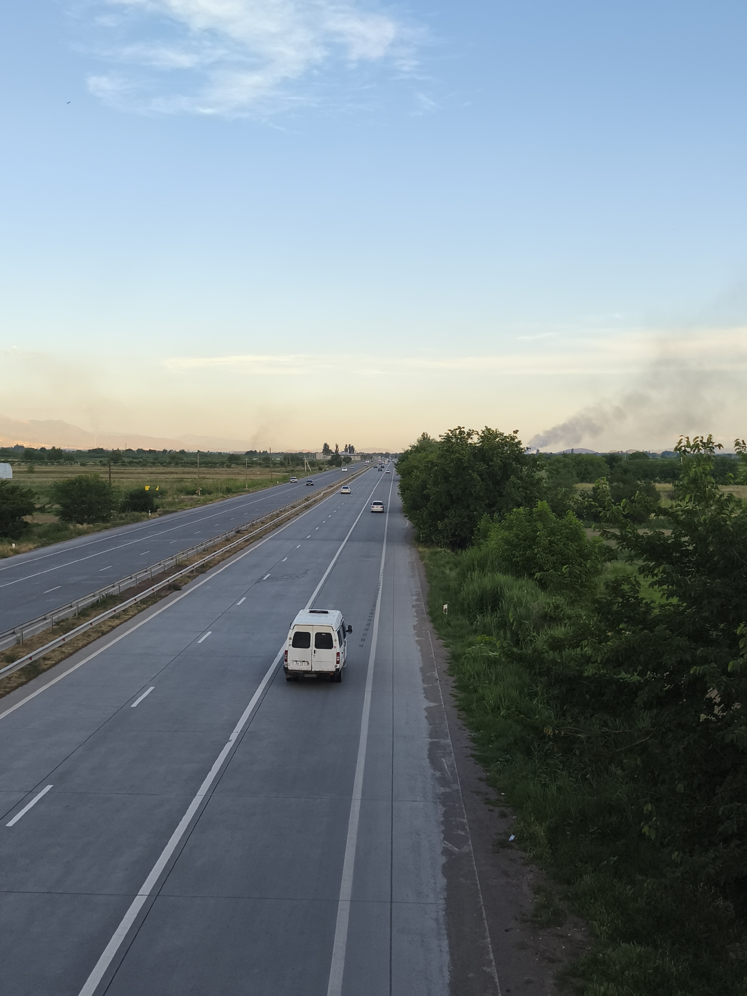 This is a high-angle shot of a highway near Azatavan, Armenia, during daylight. The sky is a pale blue with scattered white clouds. The highway itself is paved and has multiple lanes, separated by white lines. On the right side of the highway, there is a dense growth of green trees and grass. In the distance, there are fields and mountains. Smoke is visible rising from the horizon on the right side of the image. A white van is driving on the highway in the foreground, and other vehicles are visible further down the road. The overall scene depicts a rural or semi-rural highway environment.