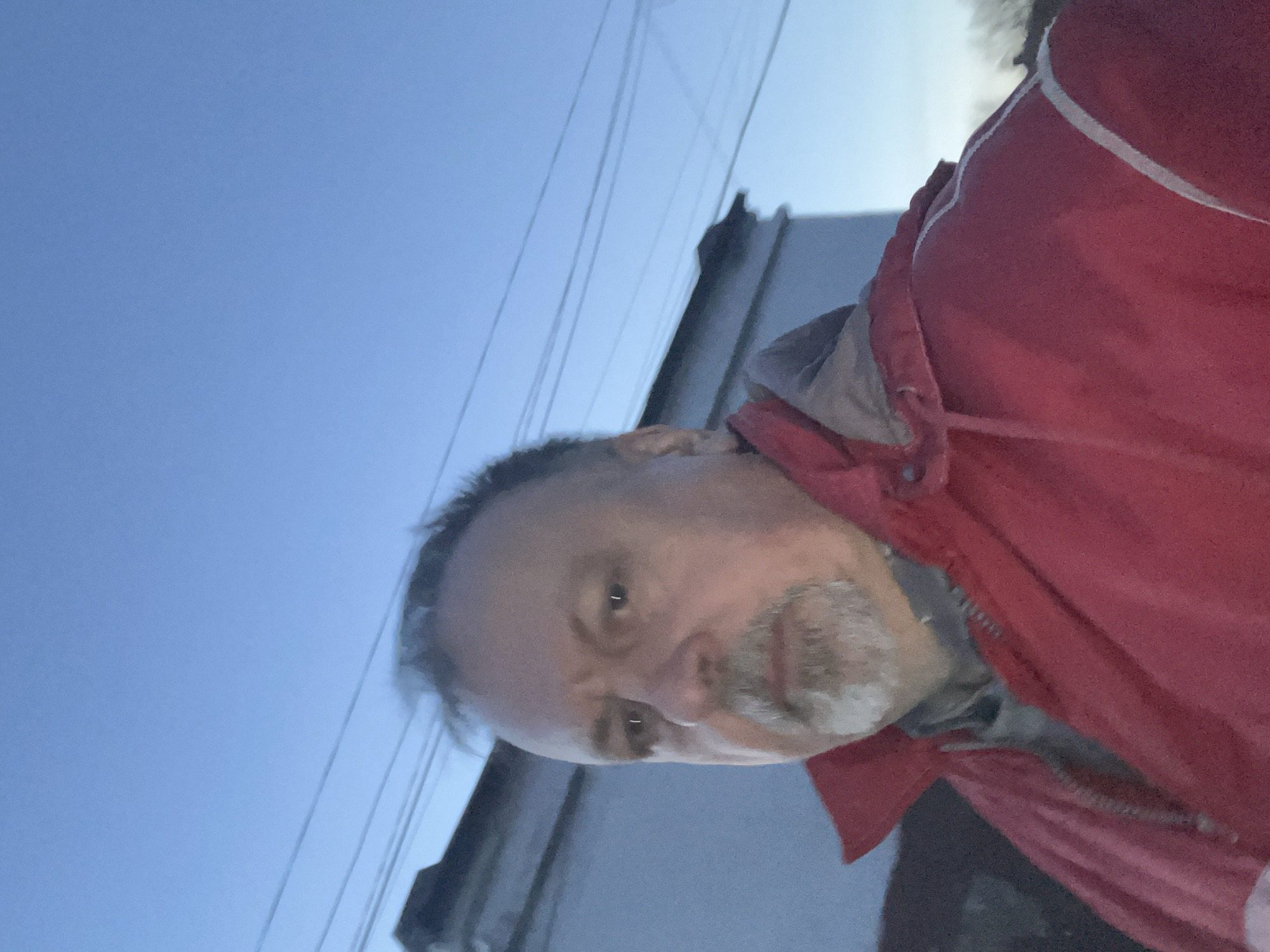A middle-aged man with graying hair and a salt-and-pepper goatee is wearing a red jacket with white trim. He is looking directly at the camera. In the background is a building and a clear blue sky with power lines. There is no information in the image to indicate the location as Podolsk, Russia.
