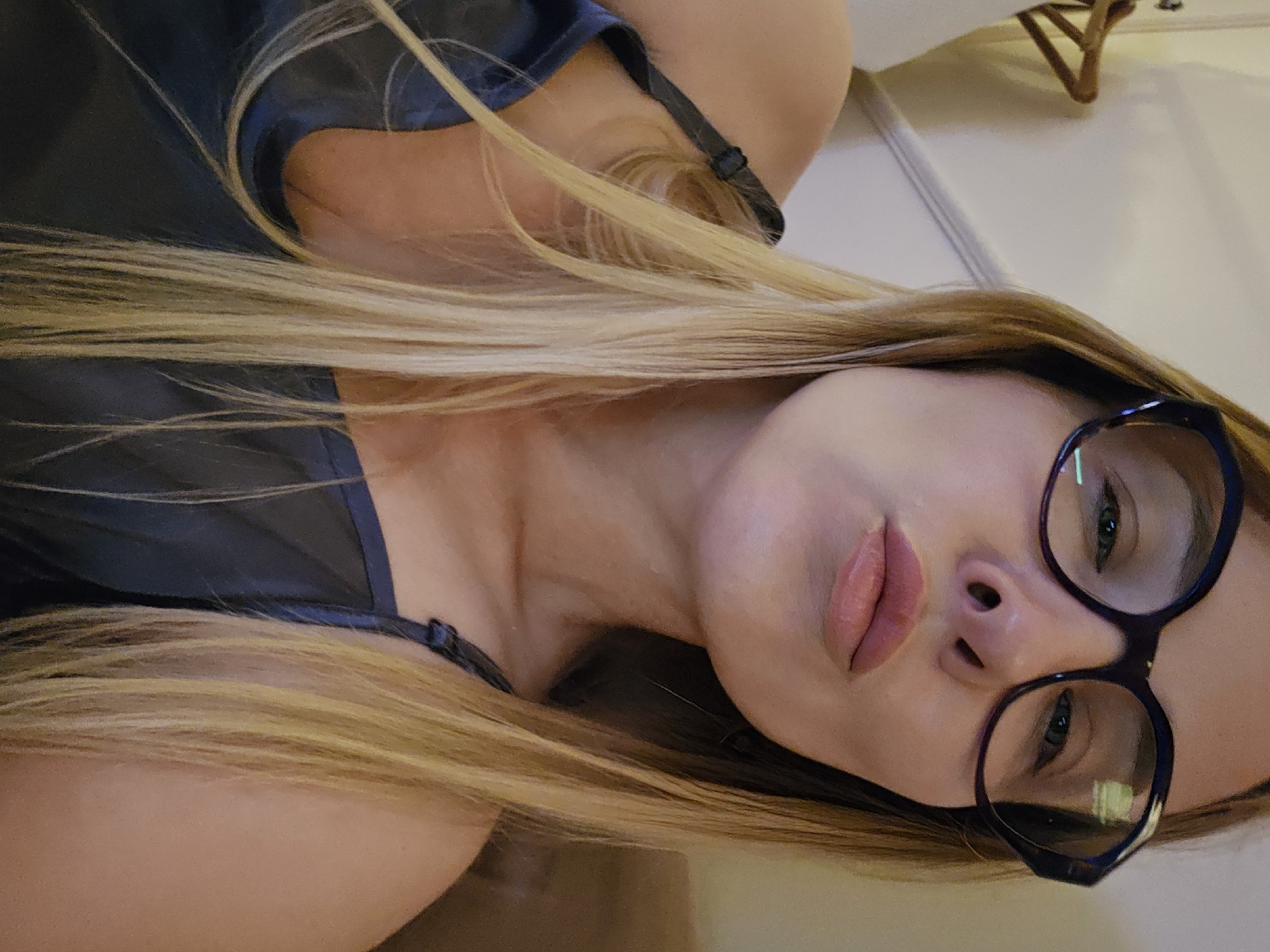 A woman with blonde hair and glasses is lying down, her head slightly tilted. She is wearing a navy blue top with thin straps. Her facial features are visible, including her eyes, nose, and lips. The background appears to be an interior setting with a white surface and a wooden object partially visible. No information about the location in Sochi, Russia is discernable from the image.