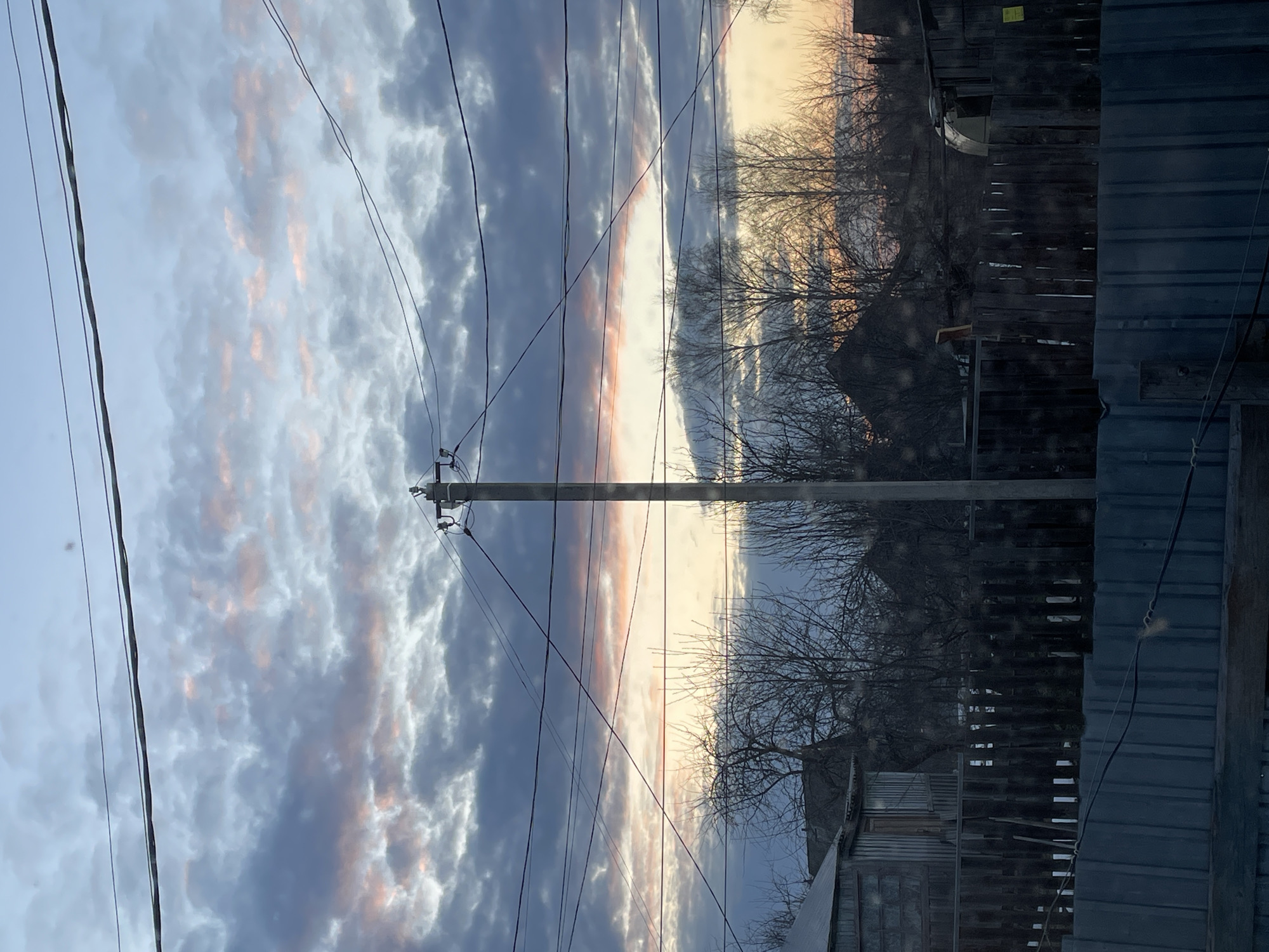 The image shows a view in Фоки, Russia. The sky is filled with clouds that are a mix of gray and white with hints of orange, suggesting either sunrise or sunset. A utility pole stands prominently in the center of the frame, with several power lines running across the sky. In the background, there are bare trees, indicating it may be winter or early spring. Below the trees are buildings and fences, possibly houses or outbuildings typical of a residential area. The photograph is taken from a position that suggests it may be behind a building or a fence itself, with parts of a structure visible in the foreground.