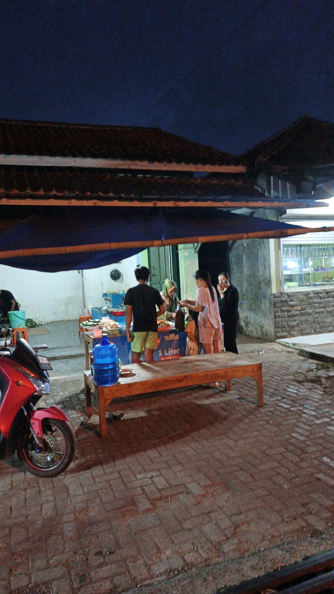 The image depicts a nighttime scene at a food stall located in Cibenda, Indonesia. The stall is set up outdoors, under a blue tarp supported by bamboo poles. A red motorcycle is parked on the left side of the frame, in front of a table displaying food items. Several people are gathered around the stall, appearing to be customers and vendors.
In the foreground, a wooden table is visible, with a large blue water jug placed on it. The ground is paved with brick-like stones. Behind the table, a man in a black t-shirt and green shorts stands facing away from the camera. To his right, a woman wearing a hijab is attending to customers. Further to the right, two women are engaged in conversation. One woman in a pinkish outfit is holding something in her hand, possibly a transaction. The other woman, in a black garment, has her hand near her face.
The background reveals more of the food stall's setup, including shelves with various items and a wall made of concrete and stone. A bright light illuminates the stall, creating shadows on the ground. The overall atmosphere suggests a busy evening at a local eatery in Cibenda.