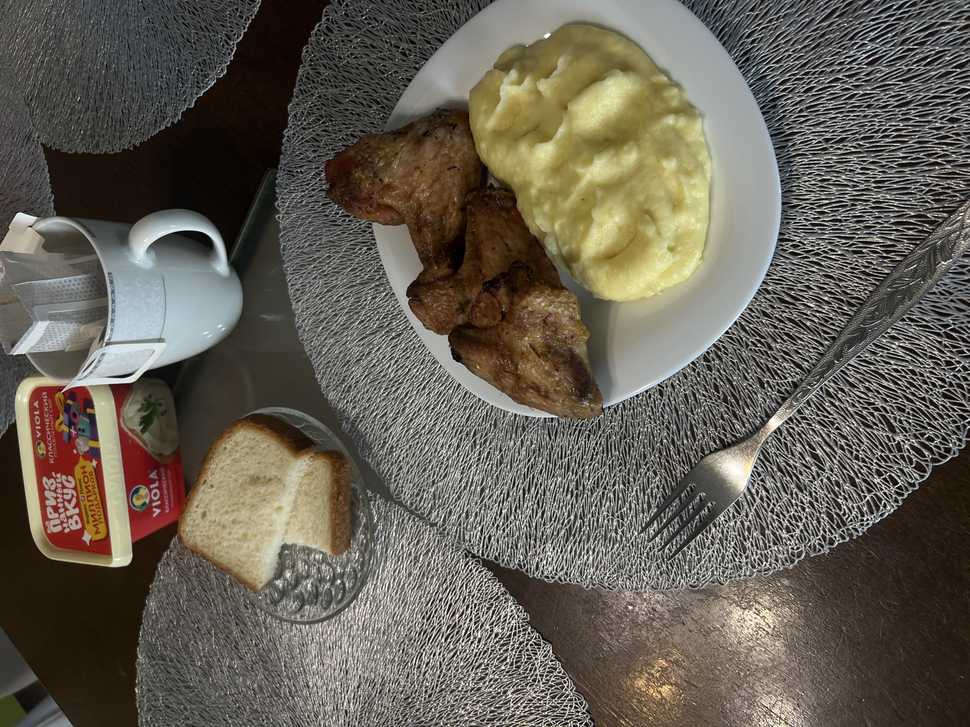 The image shows a meal served on a table. The meal includes cooked chicken, mashed potatoes on a white plate, bread in a small glass dish, and a container of Viola brand cheese spread. There's also a white mug with coffee filters inside and a fork placed near the plate. The table is covered with a metallic gray placemat. The items are arranged on a dark wooden table. The scene appears to be indoors, possibly a home or a restaurant. The container of Viola cheese spread suggests the location may be Russia. Given that "ПРИЗ НЯНЧИН" is likely a product name or promotional message, and the brand "VIOLA" is known in Russia, it's plausible the scene is in Krasnoyarsk, Russia. There are no people visible in the frame, so no actions or interactions can be directly observed, and the context is simply a meal setting.