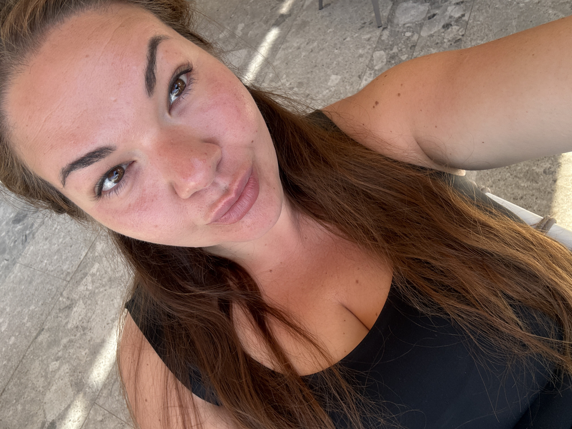 A selfie of a woman with light skin, brown eyes, and brown hair, wearing a black top. The woman is outdoors, likely in Herceg Novi, Montenegro, given the marble-like flooring that is a common feature in the city's public spaces. Sunlight and shadow indicate an outdoor setting, possibly on a terrace or patio. The woman is holding the camera at an angle, capturing herself from slightly above.