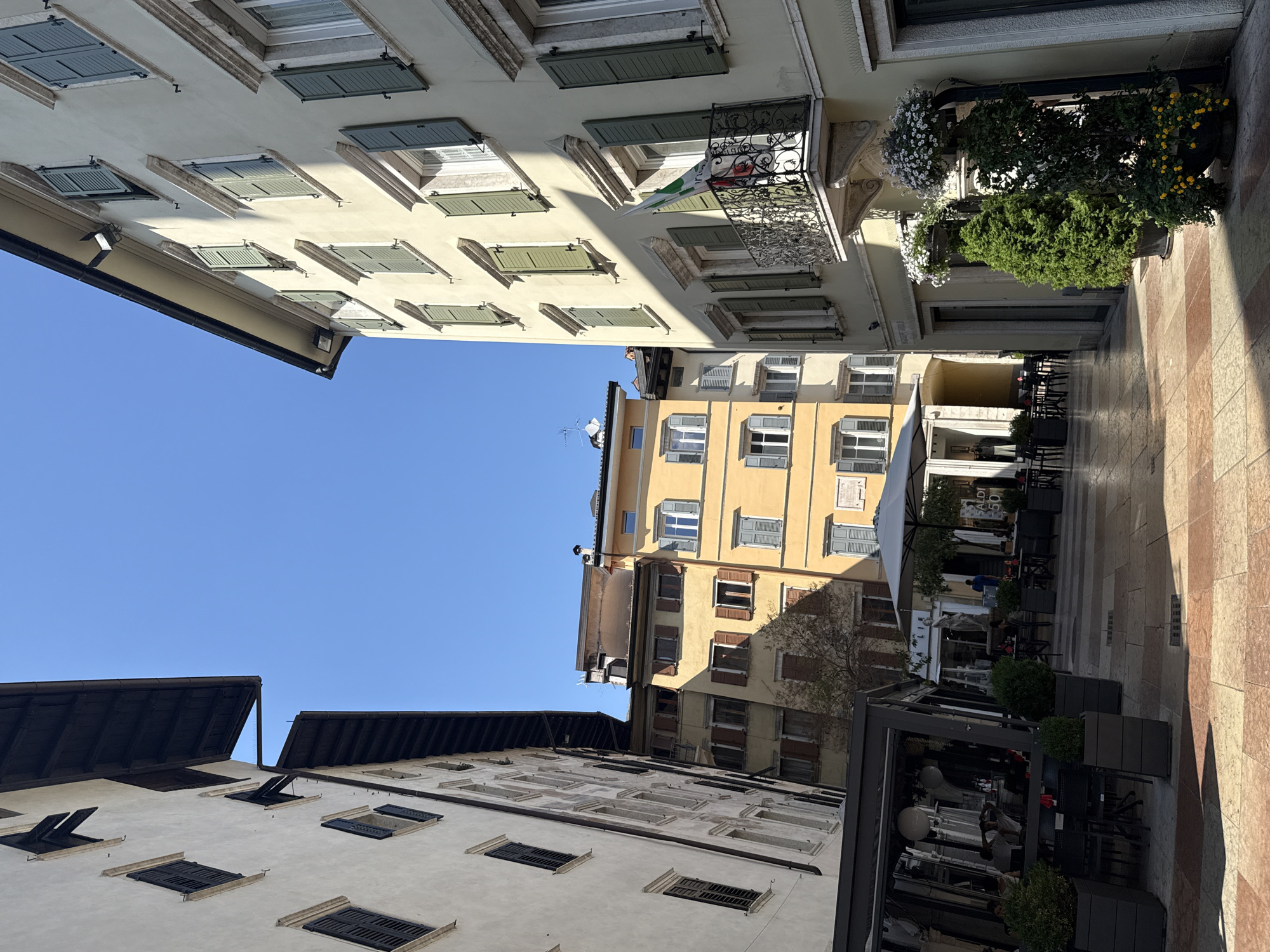 The image captures a sun-drenched street scene in Trento, Italy. On the right, a pale yellow building with green shutters dominates the foreground, featuring a decorative balcony with an Italian flag. To the left, a taller, off-white building with dark eaves lines the street, its facade punctuated by evenly spaced windows. In the background, a narrow alley opens up to reveal more buildings, including a taller yellow structure with grey shutters and a darker building with brown shutters. The street pavement is a mosaic of light and dark square tiles.
In the mid-ground, a covered outdoor seating area of a restaurant is visible, with tables and chairs set up. People are seated at some of the tables, engaged in conversation or dining. Another open area with tables and chairs is visible further down the street, shaded by a large grey umbrella. Lush green potted plants and trees are strategically placed throughout the scene, adding a touch of nature to the urban environment. The sky is a clear, bright blue.