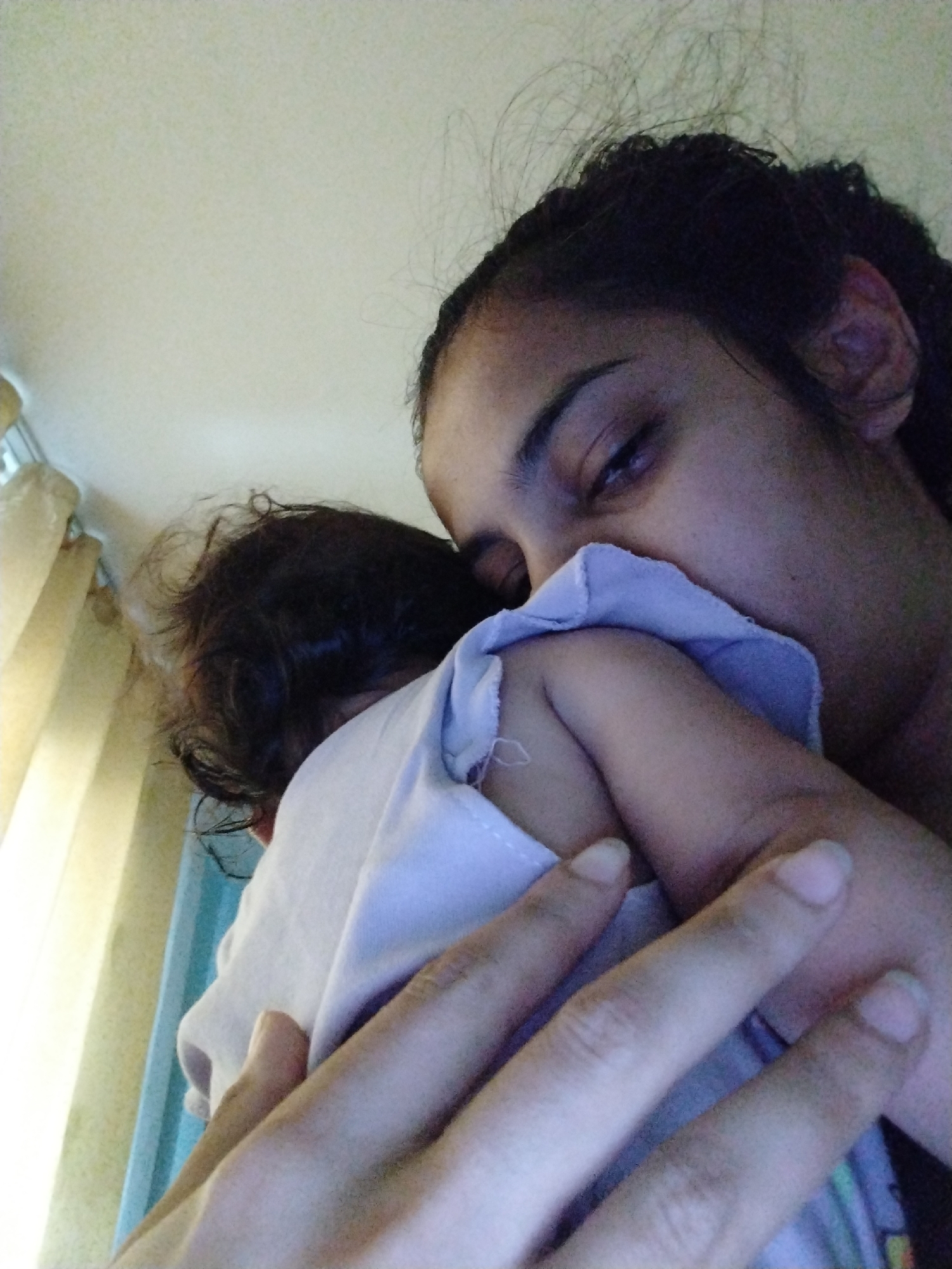 A woman is holding a baby. The woman is facing the camera and is looking down at the baby with her eyes closed. The baby is facing away from the camera and appears to be asleep. The woman is wearing a light blue shirt, and the baby is wrapped in a light blue blanket. The background is a wall and a window with blinds. The scene appears to be indoors.
