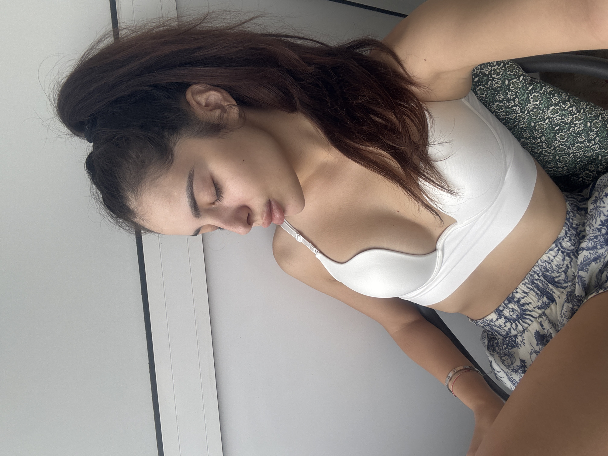 An indoor, slightly rotated, full-body shot captures a young woman resting with her eyes closed, giving an impression of peace and relaxation. Her head is tilted to her right, and her long, dark brown hair is pulled back into a high ponytail, with loose strands framing her face. She is wearing a white, seamless bralette or sports bra and blue and white patterned shorts, which appear to have a floral or toile de Jouy design. On her left wrist, two thin bracelets are visible, one in a metallic tone with small accents and another with red or pink details.
The woman appears to be reclining or leaning back, supported by a dark green patterned fabric, possibly a cushion or chair cover, and a dark chair frame visible behind her upper body. The background is a plain, light grey or off-white wall, featuring a distinct dark vertical line that could be a door frame or architectural feature. The lighting is soft and even, consistent with an indoor setting during the daytime. There is no visible text in the image. This scene is set in Plovdiv, Bulgaria.