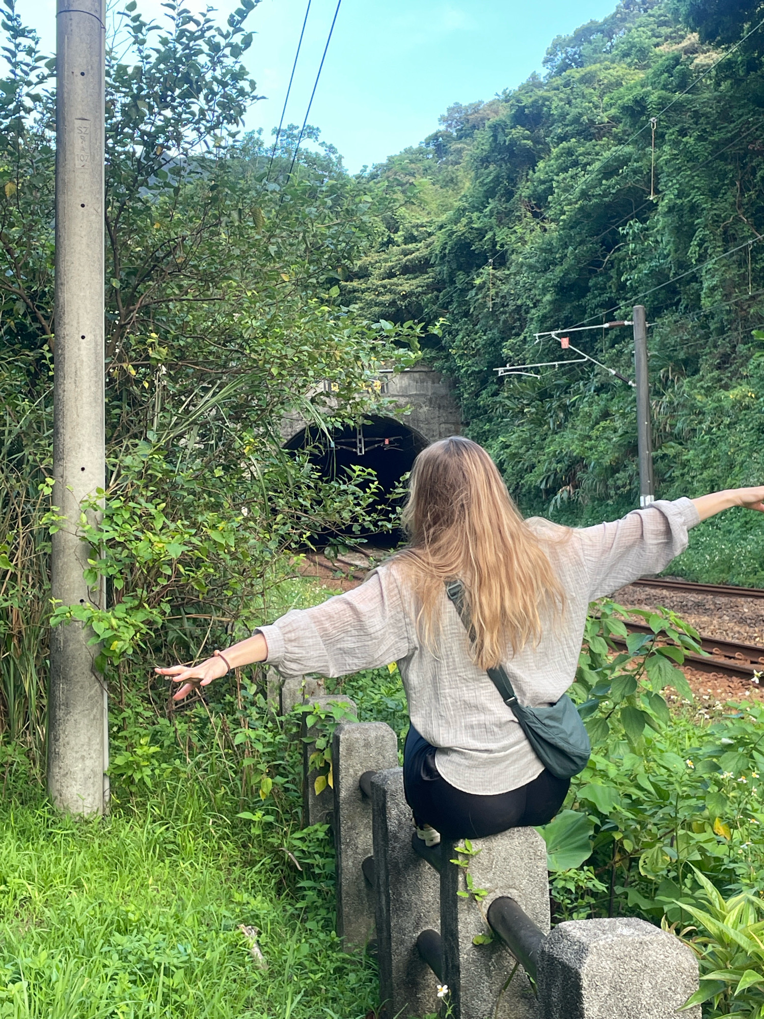 A woman with long blonde hair is sitting on a concrete fence with her back to the camera, facing a train tunnel. She is wearing a light-colored, loose-fitting shirt and dark pants, with a dark green crossbody bag slung over her shoulder. Her arms are outstretched to her sides, as if embracing the environment. The tunnel entrance is dark, with visible train tracks leading into it. The surrounding area is lush and green, with dense vegetation covering the hillside. A concrete utility pole stands to the left of the frame, and power lines are visible against the sky. The scene appears to be outdoors, possibly in a rural or semi-rural area of New Taipei City, Taiwan, given the presence of a train tunnel and the verdant landscape. The ground is covered with grass and some small white flowers.
