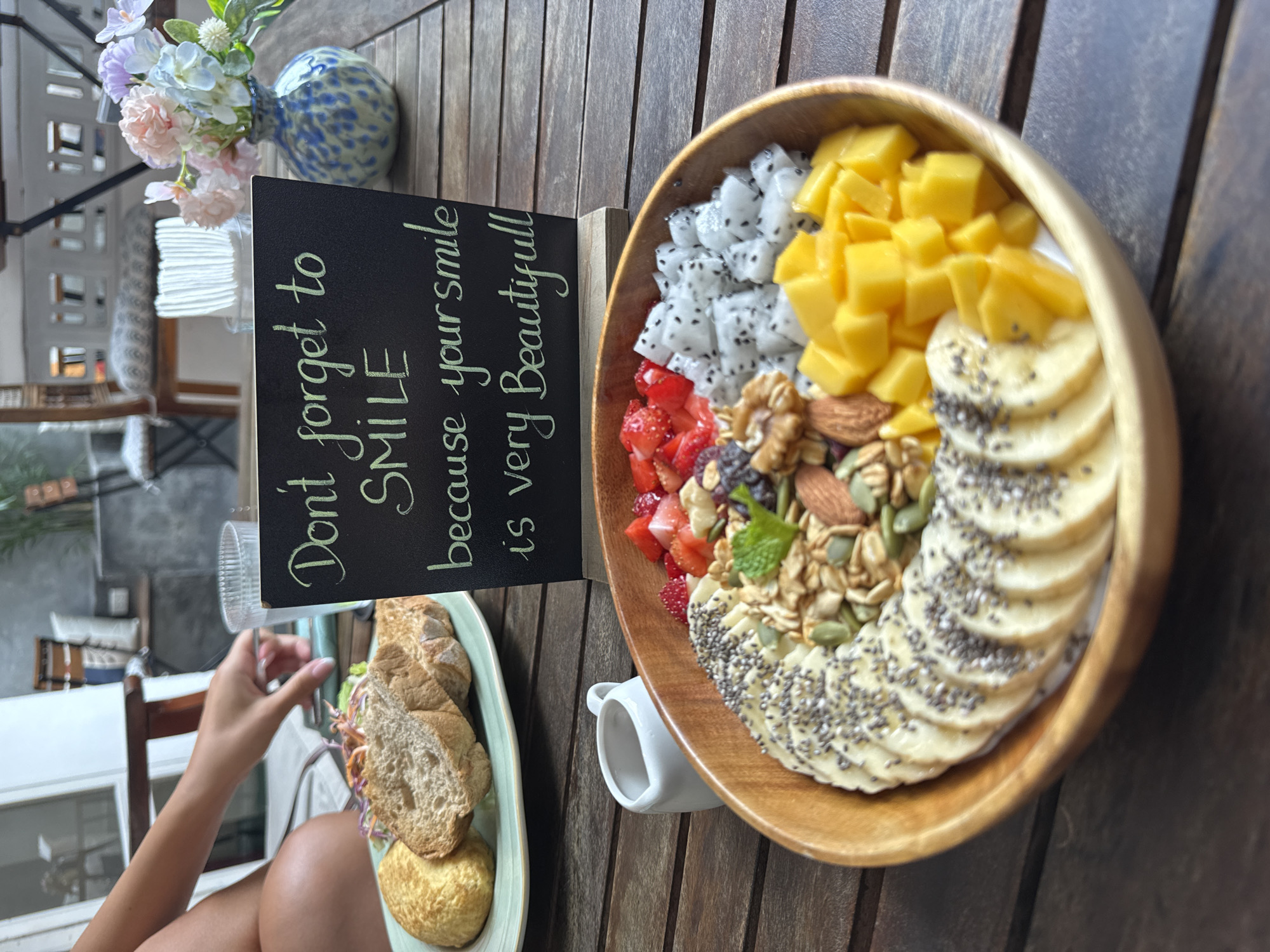The image depicts a wooden table at an outdoor cafe. On the table are a wooden bowl filled with fruit, granola, and nuts, and a plate of bread with a side salad and a bread roll. A chalkboard sign is propped up next to the bowl. It reads, "Don't forget to smile because your smile is very Beautiful!" To the left of the bowl is a small white creamer or condiment dish. In the background, there is a partially visible person holding a glass and a vase of flowers. The scene is well-lit and appears to be taken during the daytime. There is no indication that the image was taken in Đà Nẵng, Vietnam.