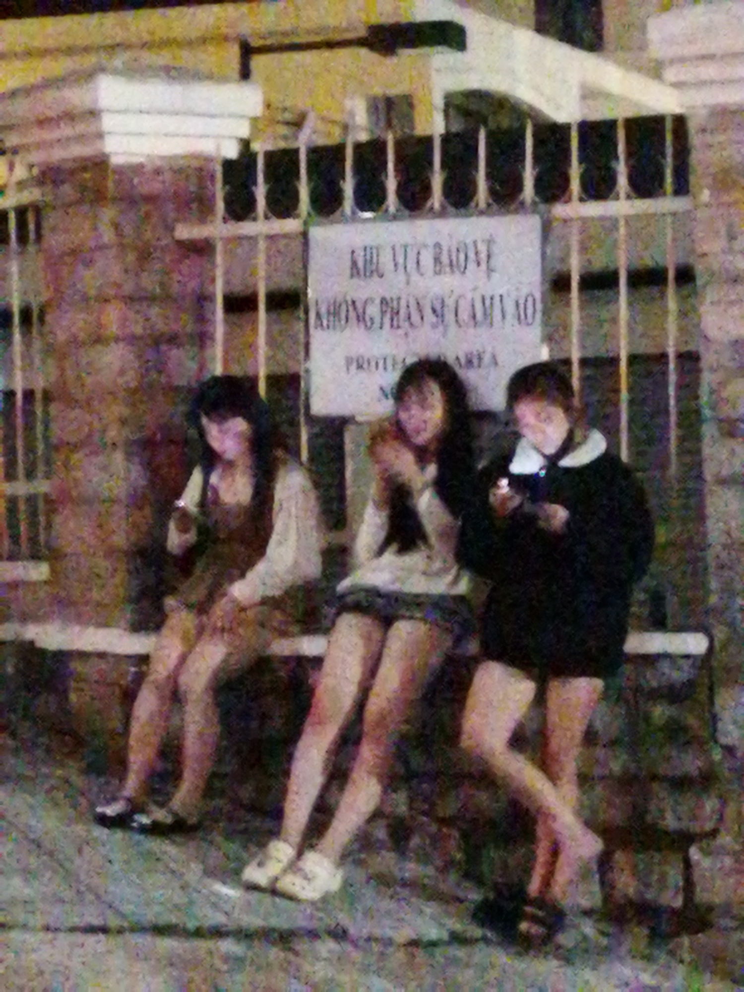 Three young women are sitting on a bench in front of a brick wall and a metal fence. They appear to be teenagers or young adults. The woman on the left is wearing a dark dress and a light-colored shirt. She is holding a small object, possibly a phone, up to her mouth. The woman in the middle is wearing a plaid skirt and a light-colored top with a dark collar. She is looking down at her phone, which she is holding in her hands. The woman on the right is wearing dark shorts and a black jacket with a white collar. She is also looking down at her phone. All three women are sitting close together, and their legs are crossed. The background is blurred and out of focus, but it appears to be an outdoor setting at night, possibly in a city or town. There is a sign behind the women with Vietnamese text. The text reads "KHU VUC BAO VE KHONG PHAN SU CAM VAO PROTECTED AREA". This translates to "Protected Area. Unauthorized personnel are forbidden to enter. Protected Area." The lighting suggests it is evening or nighttime. The overall mood is casual and relaxed, with the women engaged with their phones.