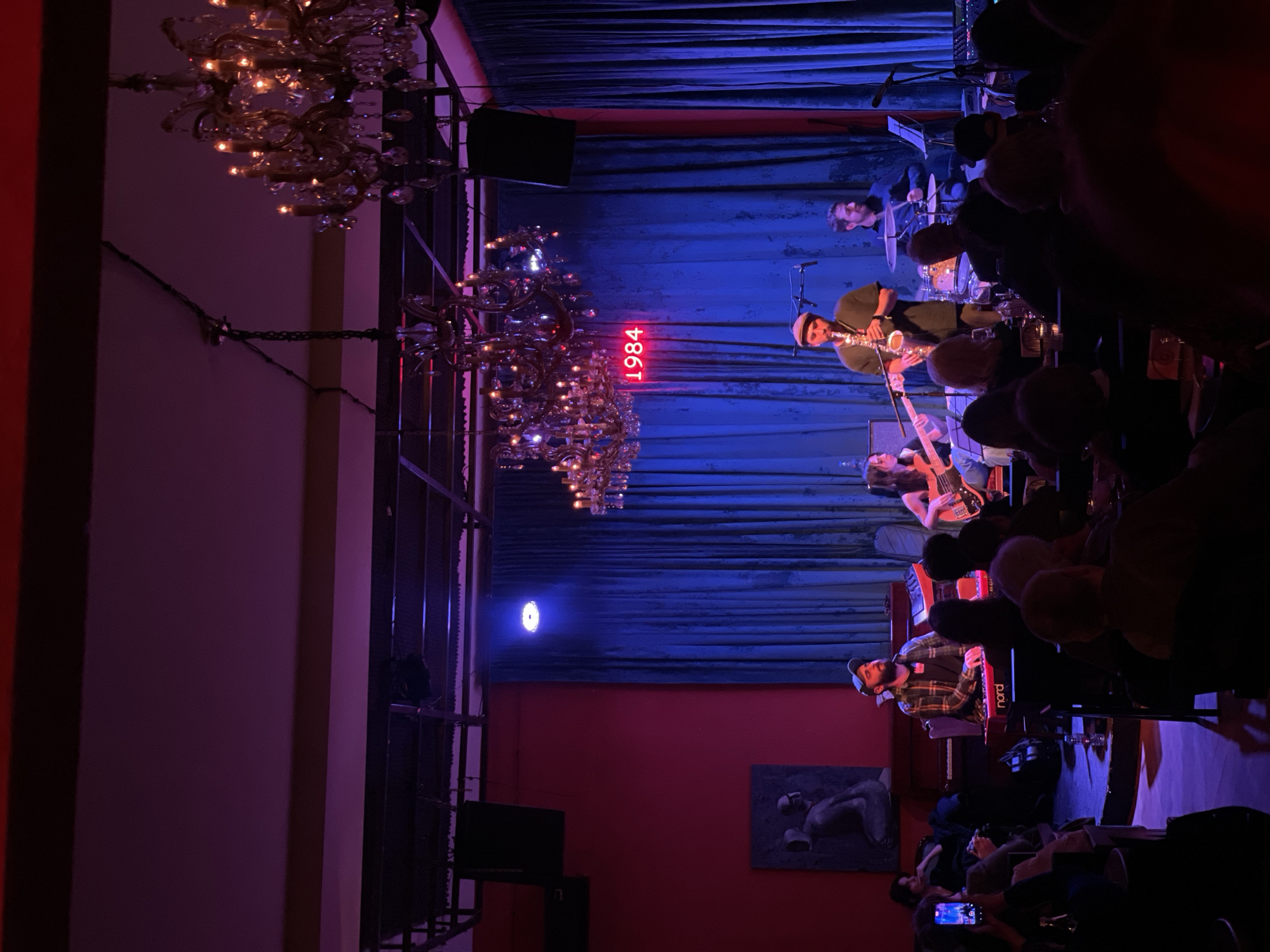 A jazz band performs on a stage in Tbilisi, Georgia. The stage is illuminated with blue and red lighting and features a backdrop of draped blue fabric. A red neon sign displays "1984". The band includes a saxophone player, drummer, bassist, and keyboardist. An audience sits at tables in the foreground, with many people appearing to be watching and listening to the performance. Chandeliers hang from the ceiling. A painting adorns the wall.