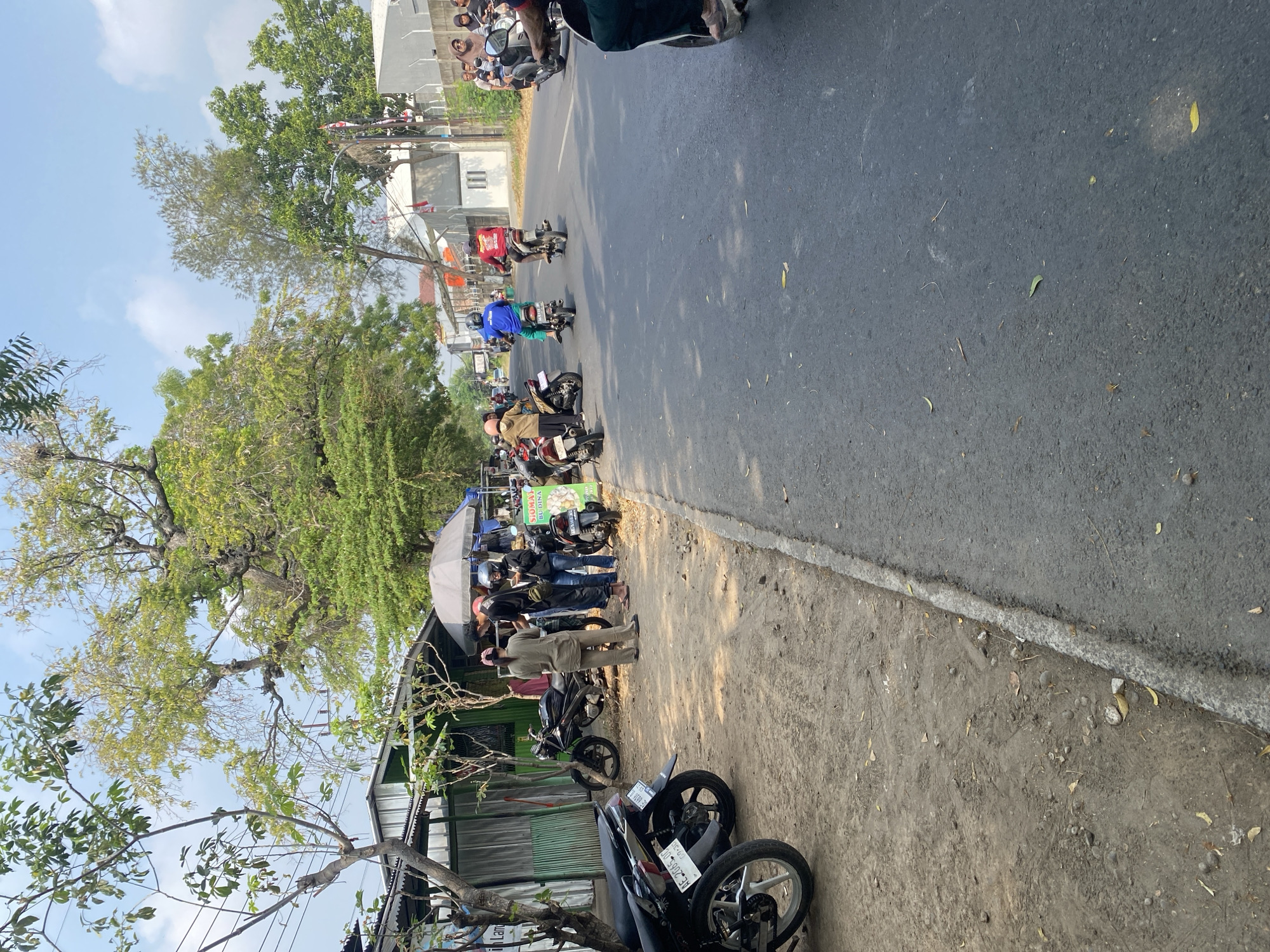 The image depicts a street scene in Banjarejo, Indonesia. Several people are gathered around a roadside food stall or cart, indicated by the presence of an umbrella and a sign. Some individuals are on motorbikes, either stopped or in motion, suggesting a busy thoroughfare. The environment includes a road with vehicles, a dirt shoulder with parked motorbikes, trees providing shade, and buildings lining the street. The sky is clear and blue. The overall impression is of a typical day in a populated area with economic activity and transportation.