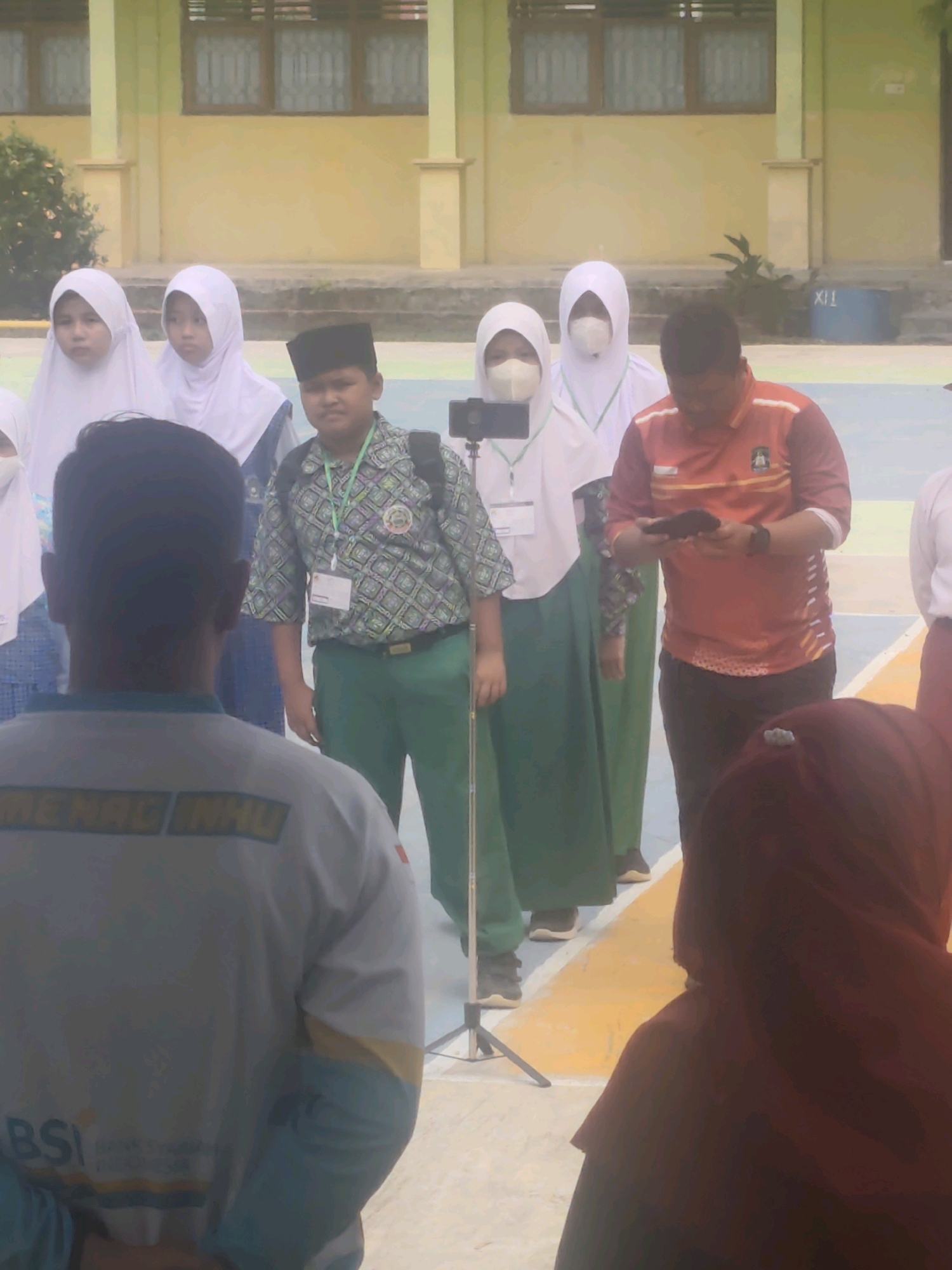 This is a vertical, medium-angle, slightly blurry photograph taken outdoors, likely at a school in Pematang Reba, Indonesia. The background features a pale yellow building with multiple dark-paned windows and visible support pillars. In front of the building, there is a gray paved area, followed by a yellow and blue patterned surface. A group of people is present, mostly facing away from the camera.
In the foreground, the back of a person wearing a light gray t-shirt with blue and yellow accents and text that appears to read "MENAG INHU" and "BSI" is prominently visible. To the right of this person, further into the scene, is a person wearing a red long-sleeved shirt with orange and white stripes, holding a dark object, possibly a phone.
Behind these individuals, a group of students, all wearing white hijabs, are standing. Some of them have light-colored masks covering their faces. One student in the center, also wearing a white hijab, is wearing a green patterned shirt with a lanyard and a black peci (a cap). To the left of this student is another student in a white hijab and a blue garment. Another student in a white hijab is visible further to the right.
A black tripod with a mobile phone mounted on it is positioned in front of the students, suggesting a recording or live-streaming activity. The overall atmosphere appears to be one of an organized event or gathering, possibly a school ceremony or educational activity.