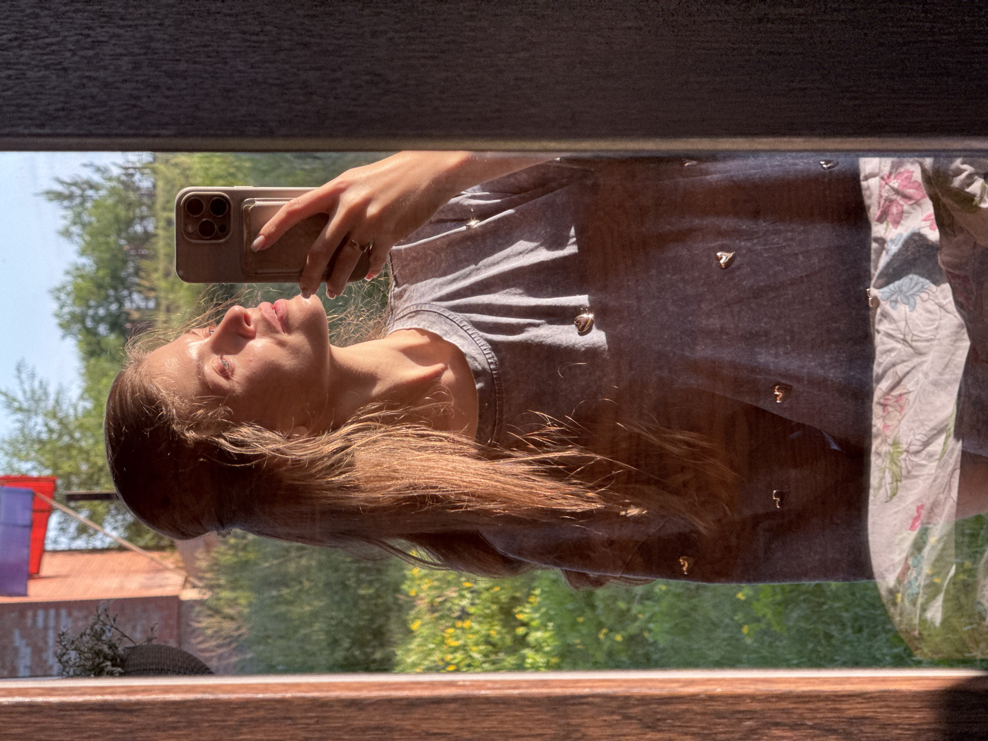 A young woman is taking a selfie in a mirror, holding a smartphone in her right hand. She has long, light brown hair and is wearing a gray t-shirt adorned with small heart-shaped embellishments. The background reflected in the mirror shows a grassy area with scattered yellow flowers and trees, suggesting an outdoor setting. In the upper left corner of the reflection, a brick building and a flag are visible, possibly indicating a location within Minusinsk, Russia. The lighting suggests it is either sunrise or sunset. The woman is wearing a ring on her left ring finger.