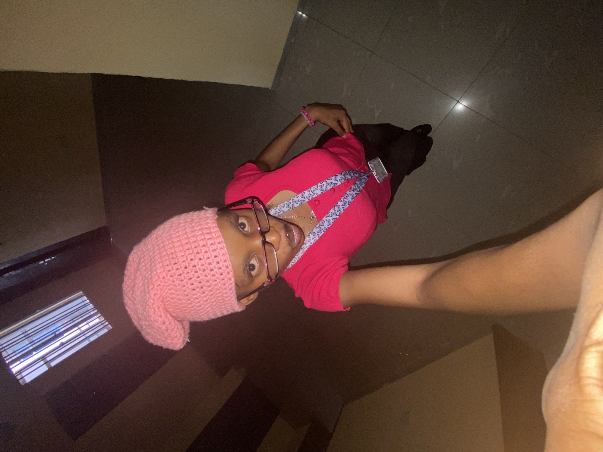 A person wearing a pink knit cap, glasses, and a pink top with a blue butterfly-patterned strap is taking a selfie. They are indoors, possibly in a hallway with dark brown walls and a tiled floor. There is a window with blinds to the left. The city and region are not specified.