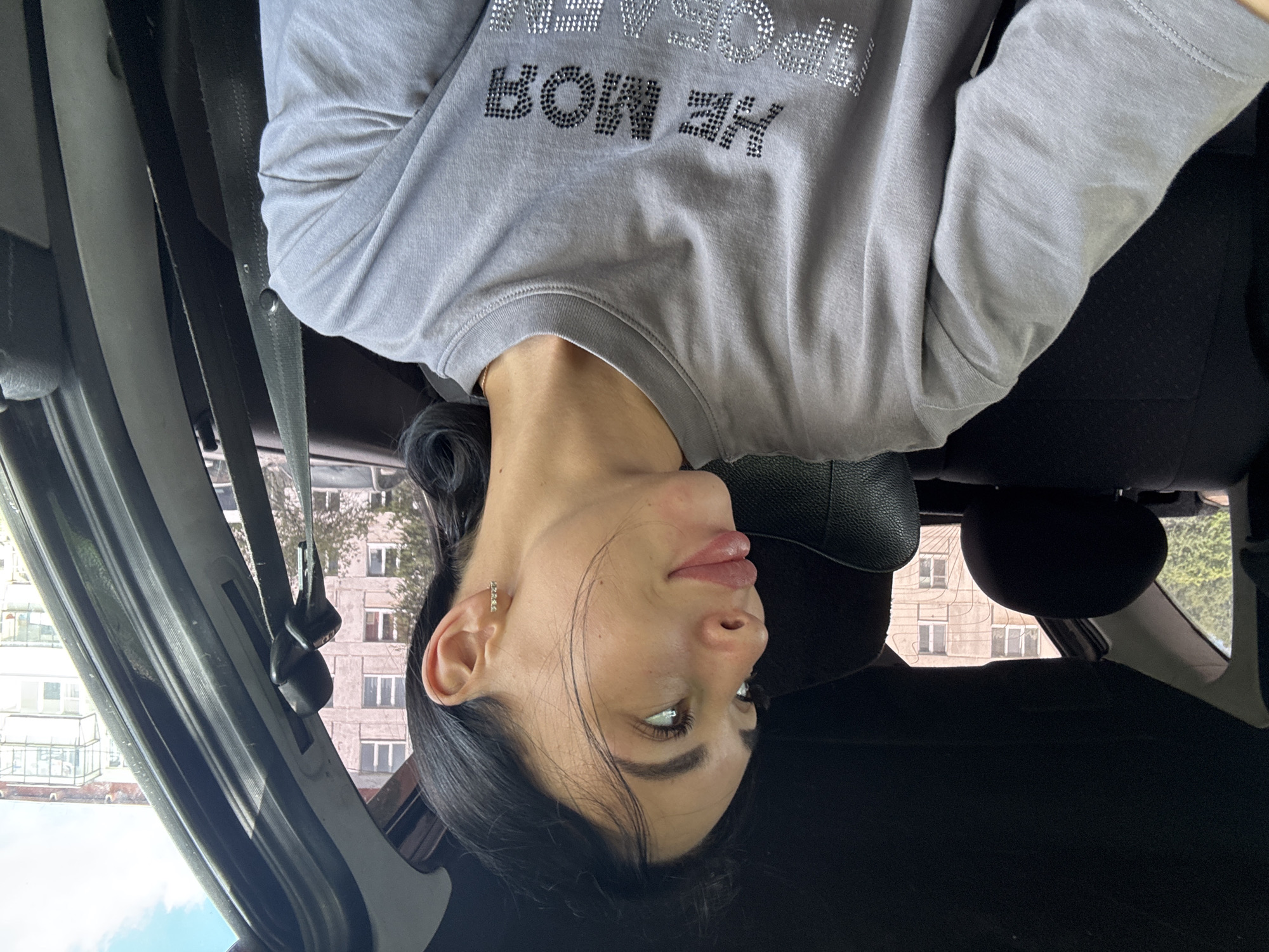 The image shows a young woman with dark hair and light skin sitting in the passenger seat of a car. She is wearing a gray t-shirt with rhinestones spelling out "HE MOЯ ПРОБЛЕМЫ" which translates to "NOT MY PROBLEMS". The woman is looking to her left, with her mouth slightly open. The background shows buildings and trees, suggesting an urban or suburban setting. The car interior is visible, including the headrest of the seat in front and the seatbelt. The woman is likely in Chaikovsky, Russia, given the context provided.