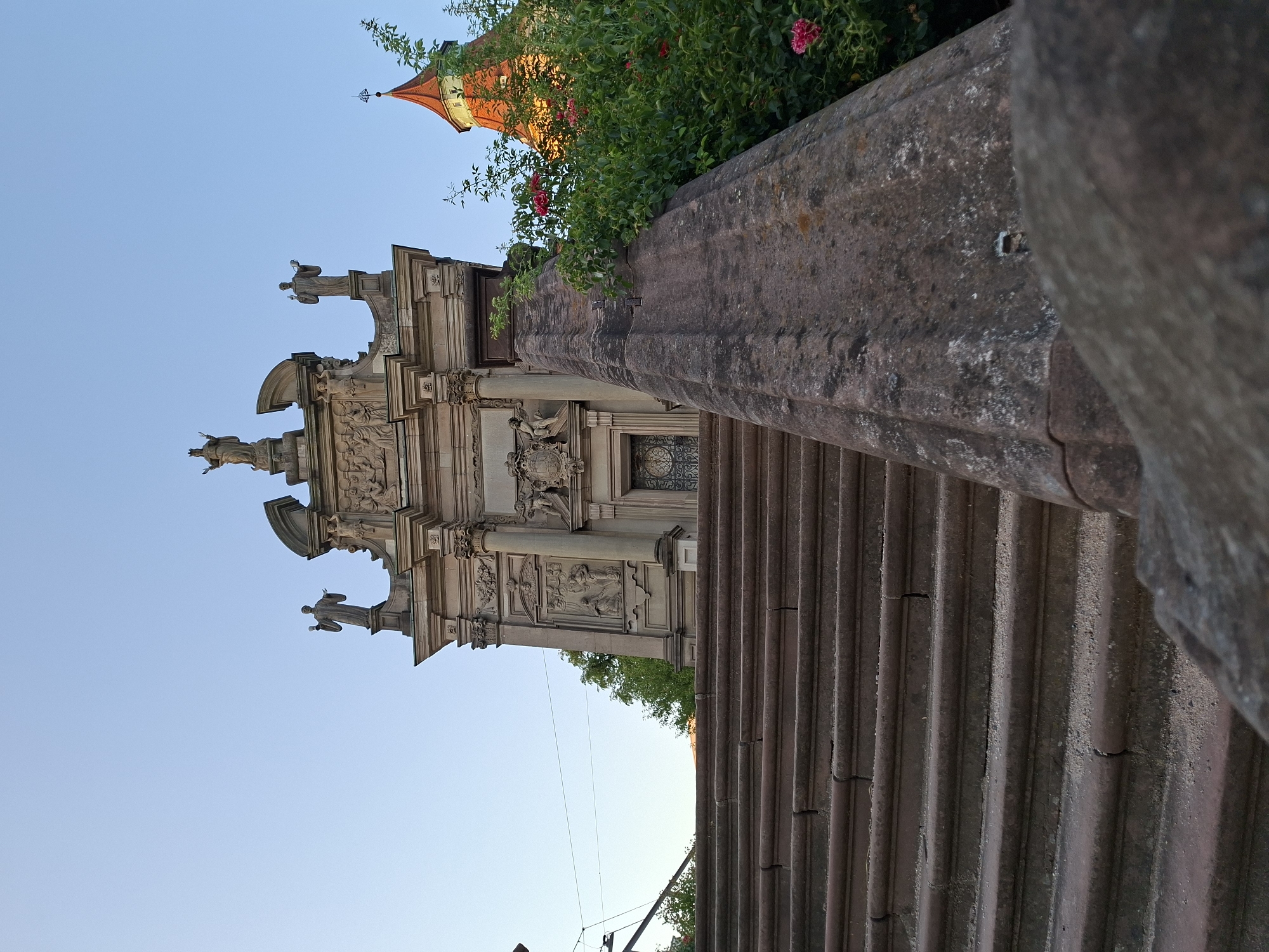 The photograph captures a low-angle view of a grand stone staircase leading up to an ornate building facade. The building appears to be constructed of stone, with intricate carvings and statues adorning its upper sections, including figures of individuals in what seem to be religious attire. To the right of the building, a distinct red-roofed tower with a spire is visible behind a lush green bush dotted with red roses. The sky above is a clear, pale blue, suggesting it is daytime, possibly late afternoon or early evening given the lighting. The foreground is dominated by the stone steps and a carved stone railing, showing signs of age and weathering. The overall scene evokes a sense of historical architecture and possibly a religious or cultural landmark. This location is the Baroque staircase leading to the pilgrimage church "Maria Hilf" in Rastatt, Germany.
