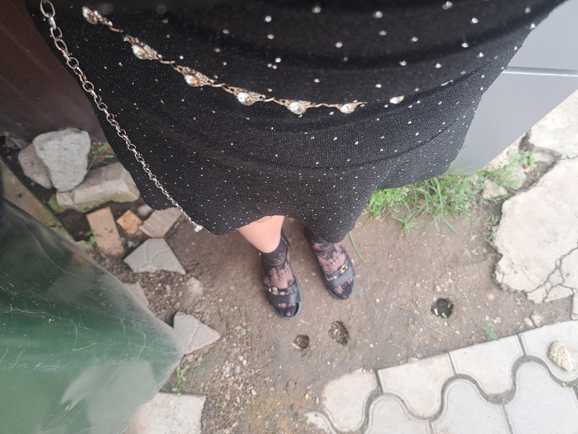 The image shows a top-down view of a person standing outdoors, likely in a yard or near a building. The person is wearing a black dress with small, light-colored dots, a thin chain belt, black patterned tights, and black sandals. The ground is a mix of cracked concrete, dirt, and paving stones. There is some grass visible along the edges. Part of a green object, possibly a container, is visible on the left. The location is Leningradskaya, Russia.