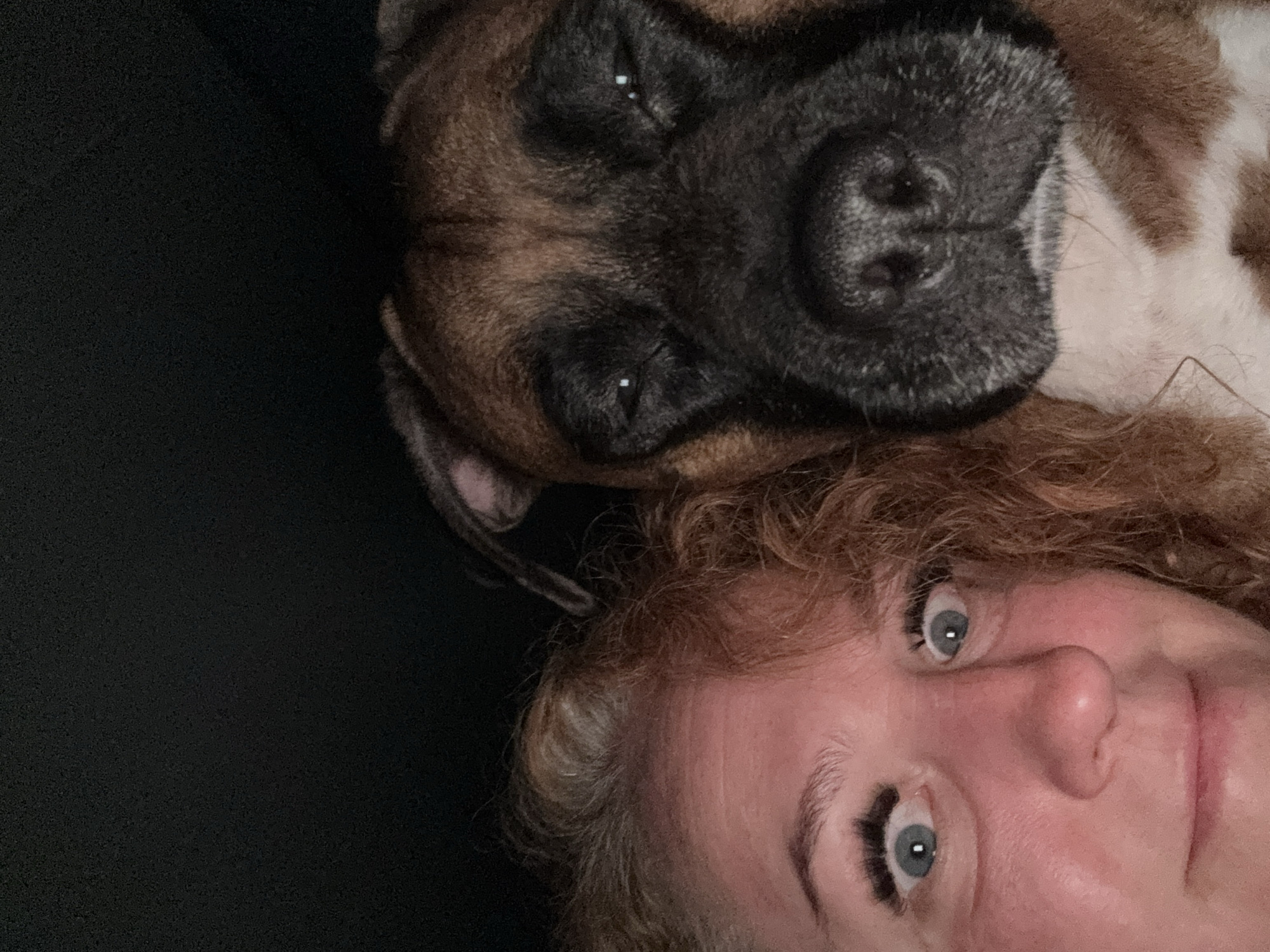 A selfie shows a woman with red curly hair and blue eyes lying down with a brown and white Boxer dog. The dog's head rests on or near the woman's head. The background is dark, possibly black fabric. The photograph is taken at a close-up angle. The location is not identifiable as Chaikovsky, Russia.