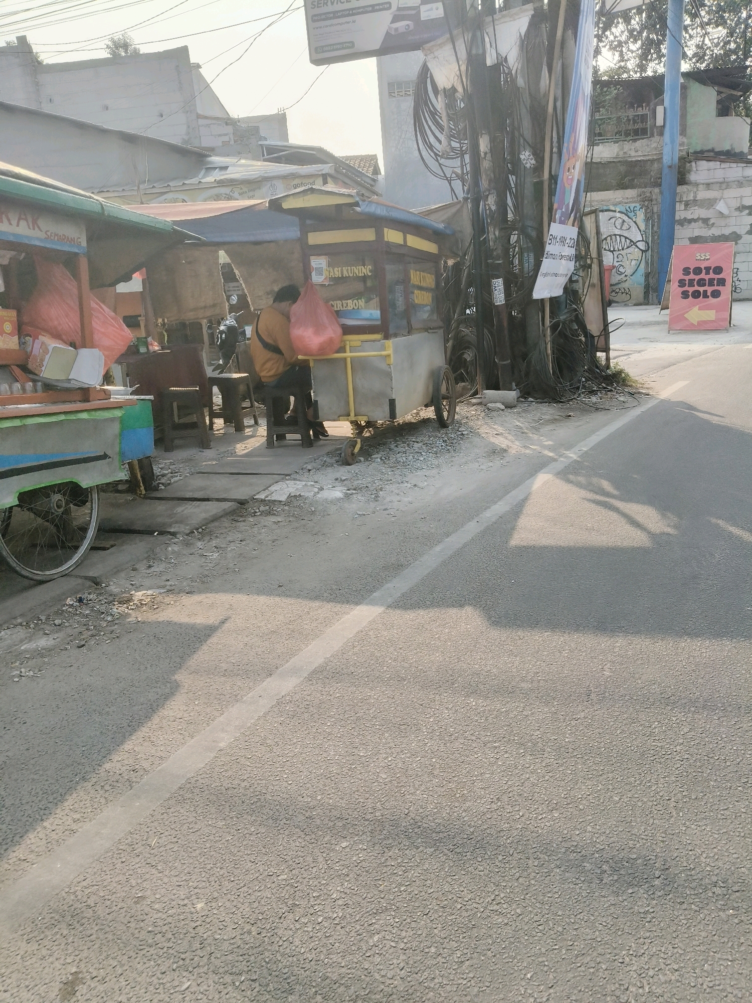 The image shows a street scene in South Tangerang, Indonesia. In the foreground, a paved road with a white dividing line runs diagonally from the bottom left to the top right. Shadows of people and objects are cast on the road surface. On the left side of the road, a food cart with a green and blue frame is parked, with a metal wheel visible. Behind it, another food cart with a yellow awning is set up. A person wearing a yellow jacket is sitting on a stool behind this cart, holding a red plastic bag. A red plastic bag is also visible on the left food cart. Behind the carts, there are several wooden stools and tables. A cluttered utility pole with numerous electrical wires and some banners is situated to the right of the food carts. One banner is white with black text and a phone number. Another banner is colorful and features a cartoon character. A red and yellow sign with text and an arrow pointing to the left is also visible near the utility pole, indicating "SOTO SEGER SOLO." The background consists of low-rise buildings, some appearing weathered, and trees. The sky is bright and hazy, suggesting a sunny day. The overall impression is that of a busy street with small food vendors.