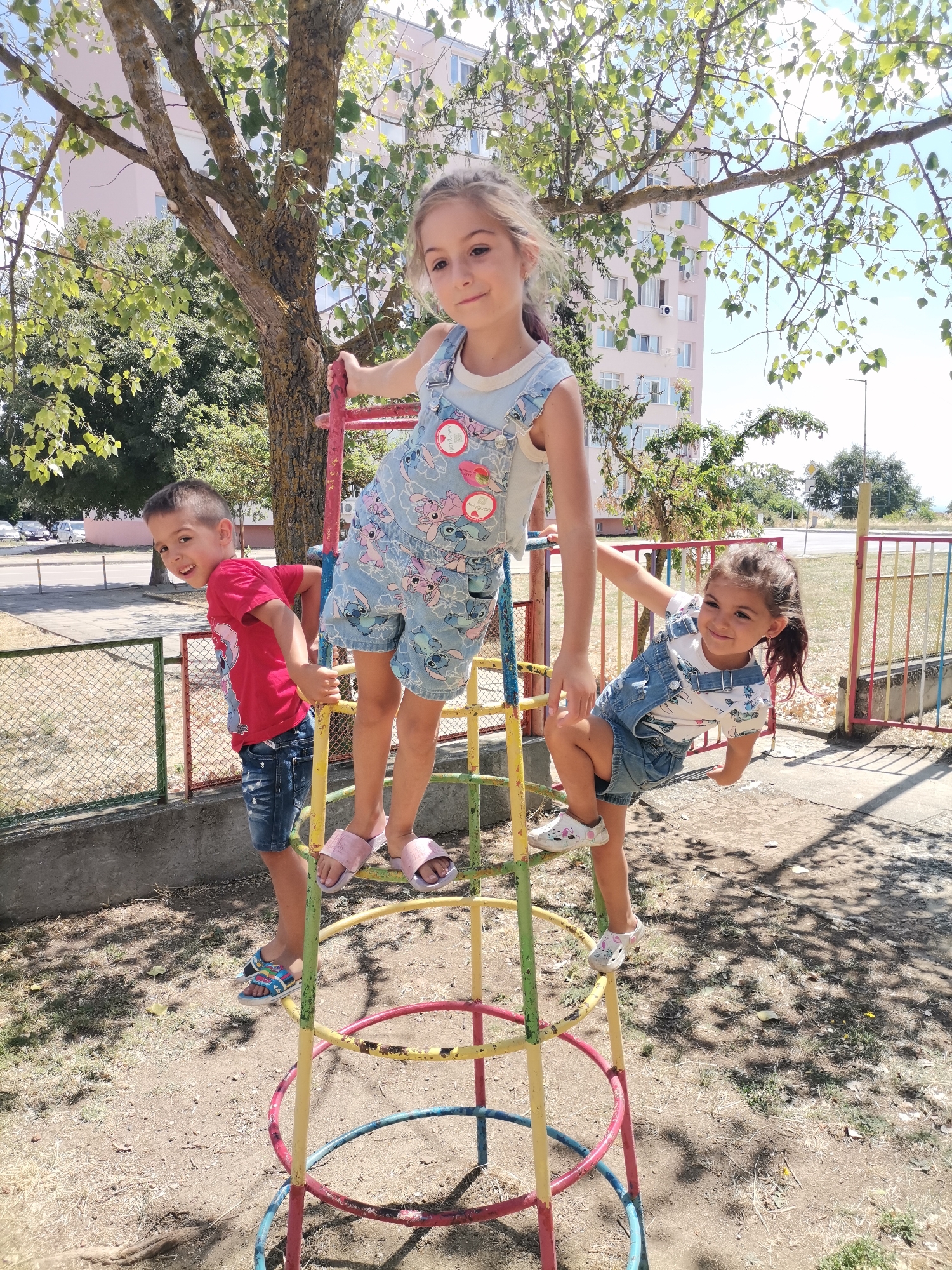 Three children, two girls and one boy, are on a circular climbing frame at a playground. The boy, wearing a red t-shirt and denim shorts, is on the left side, looking at the camera and smiling. The older girl, in blue overalls with a cartoon print, is standing in the middle, also looking at the camera. The younger girl, in a denim overall dress, is on the right, facing away from the camera. The playground is situated in a residential area of Balchik, Bulgaria, with a tall, light-colored apartment building in the background. The ground is covered with sand and dry grass, and the climbing frame is colorful with red, yellow, green, and blue sections. Trees with green leaves are scattered around, and a bright blue sky is visible. The scene appears to be on a sunny day.